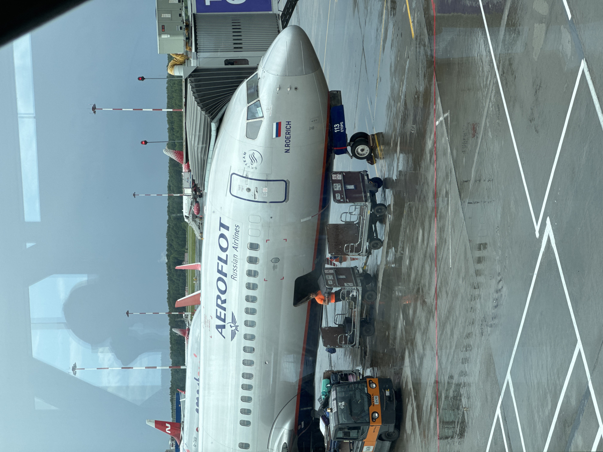 This image captures an Aeroflot Russian Airlines aircraft parked at a jet bridge in St. Petersburg, Russia. The scene is set on a wet tarmac, indicating recent or ongoing rain. In the foreground, a baggage tug with the identification "STILL 1630" is visible, along with several baggage carts. A worker in an orange shirt is attending to the cargo loading process. The aircraft, a Boeing 737 with the name "N. ROERICH" painted on its fuselage, is connected to the jet bridge. Other aircraft tails, featuring the "N" logo of Nordwind Airlines and the red and white pattern of Rossiya Airlines, are visible in the background, along with airport lighting poles. The sky is overcast and grey, suggesting a potentially stormy or rainy day.