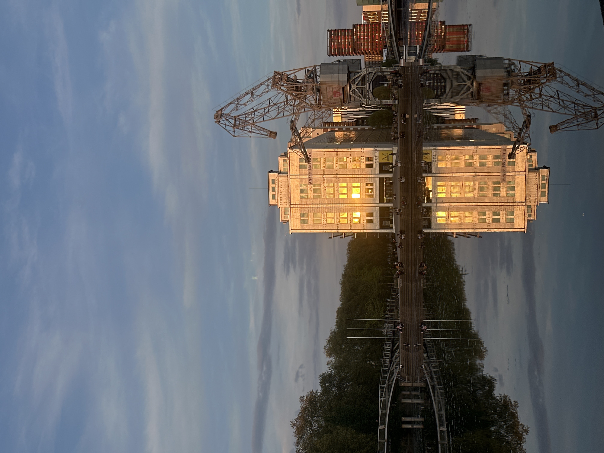 This is a photograph of the Musée d'Art Moderne et Contemporain (MAMCS) in Strasbourg, France, reflected in the water. The museum building is a multi-story structure with numerous windows that are reflecting sunlight, giving them a bright appearance. To the side of the building, there is a large, old crane structure. A pedestrian bridge with people walking on it is visible in the lower portion of the photograph. The sky is partly cloudy. The overall lighting suggests it is either sunrise or sunset.