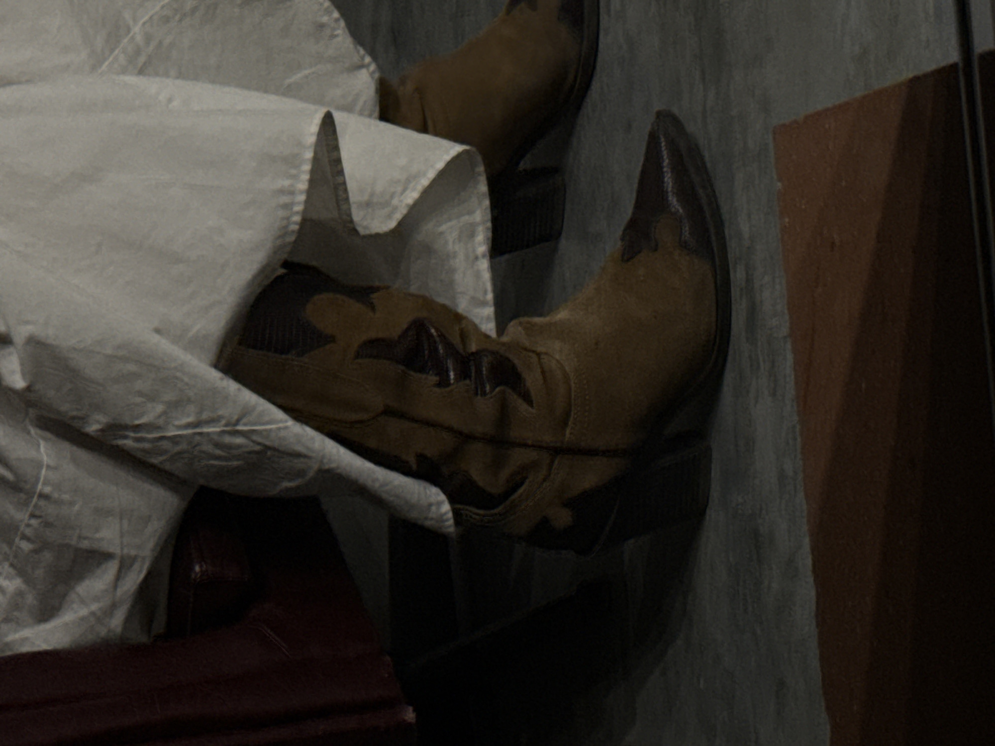 The image depicts a pair of cowboy boots. One boot is mostly visible, featuring brown suede with darker brown leather accents. A partially visible white garment drapes over the boot and part of a burgundy leather chair. The other boot is partially visible further back. The background consists of a gray surface and a brown wooden object. The image's location cannot be determined from the visual information provided.