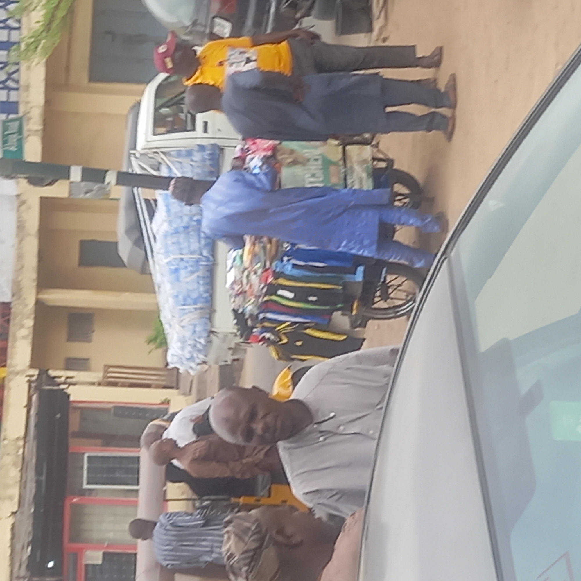The scene depicts a street in Dadi Riba, Nigeria. Several individuals are visible, including men dressed in casual attire and traditional blue robes. One man is pushing a cart loaded with bottled water and colorful clothing. The background features a building with windows and a nearby sign. The image appears to be taken from inside a vehicle, with part of the car's interior visible. The environment suggests a typical urban scene with commercial activity.