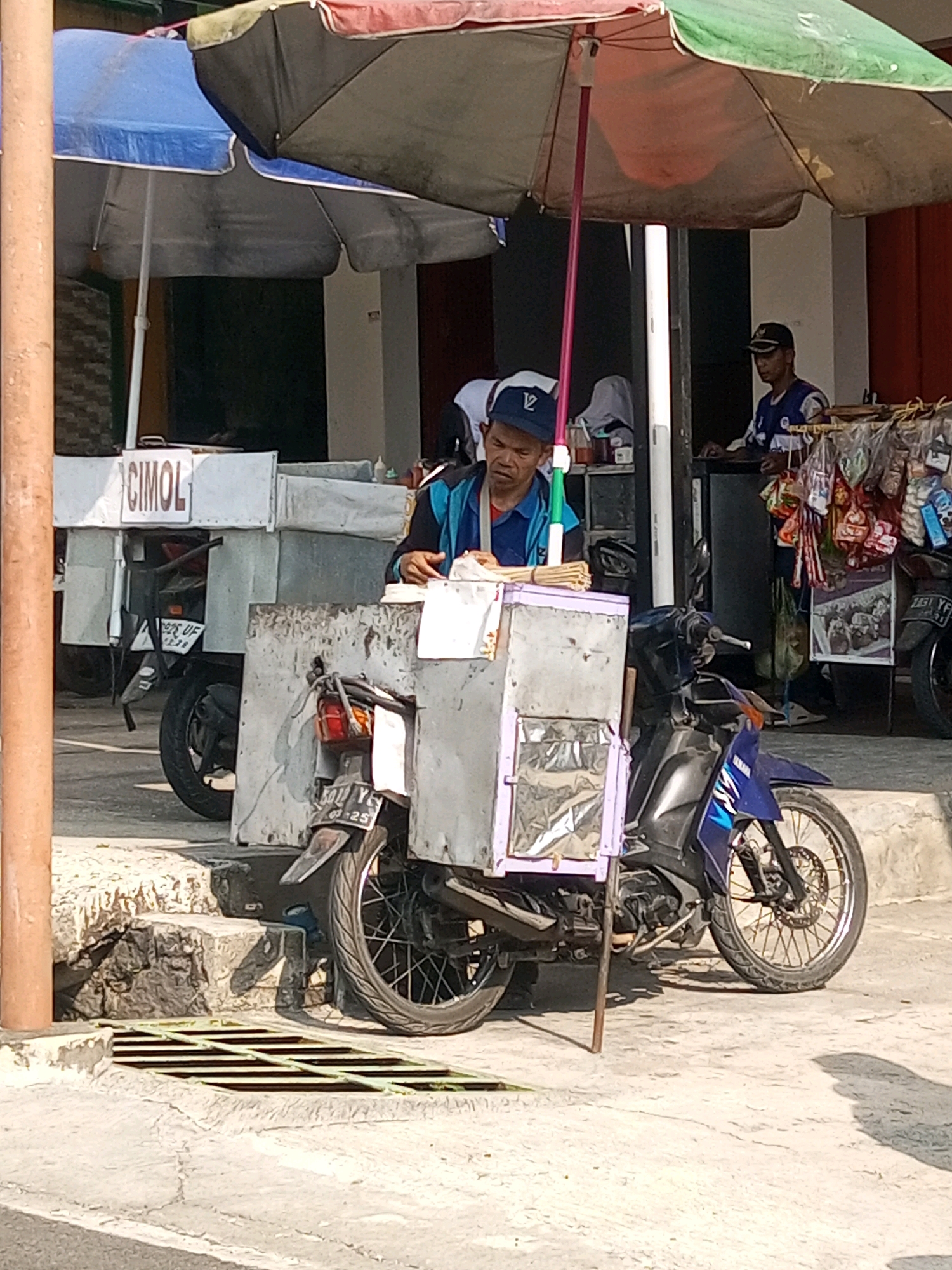 In the city of Krasak, Indonesia, a street vendor operates a food cart mounted on a motorcycle. The vendor, a man wearing a blue shirt and a dark baseball cap, is standing behind the cart, looking down. He is in the process of serving food or preparing an order, with a pile of skewers on the cart in front of him. The food cart itself is a metallic box with a purple-tinted window, attached to the back of a blue and black motorcycle. Another motorcycle is parked behind the food cart, and its license plate is partially visible, reading "BF 3378 A". In the background, a second vendor is visible, wearing a dark blue vest and a cap, standing in front of a stall with various snacks and drinks displayed. The scene is outdoors, with umbrellas providing shade over the vendor and the food cart. The ground is paved with concrete, and a metal grate is visible on the left side of the image. The overall atmosphere suggests a busy street market or food stall area.