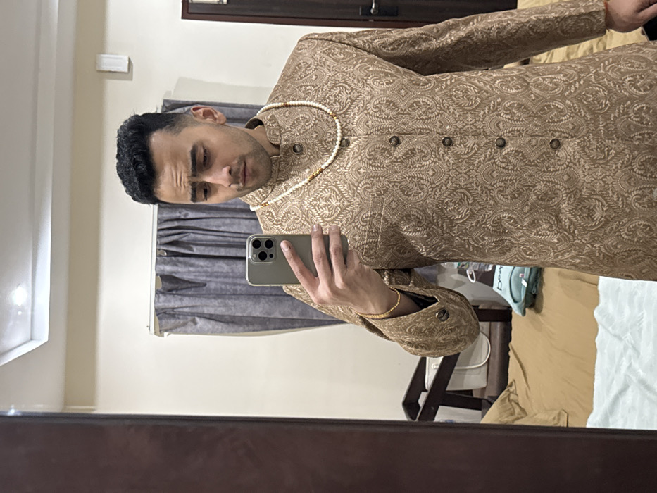 A man is taking a mirror selfie. He wears a tan sherwani with an intricate paisley pattern and a white pearl necklace. He holds a smartphone in his right hand. In the background, curtains hang by a window, a wooden door frame is visible at the top and the mirror frame is visible on the bottom. Bedding and a chair can be seen in the reflection to the right of the man.