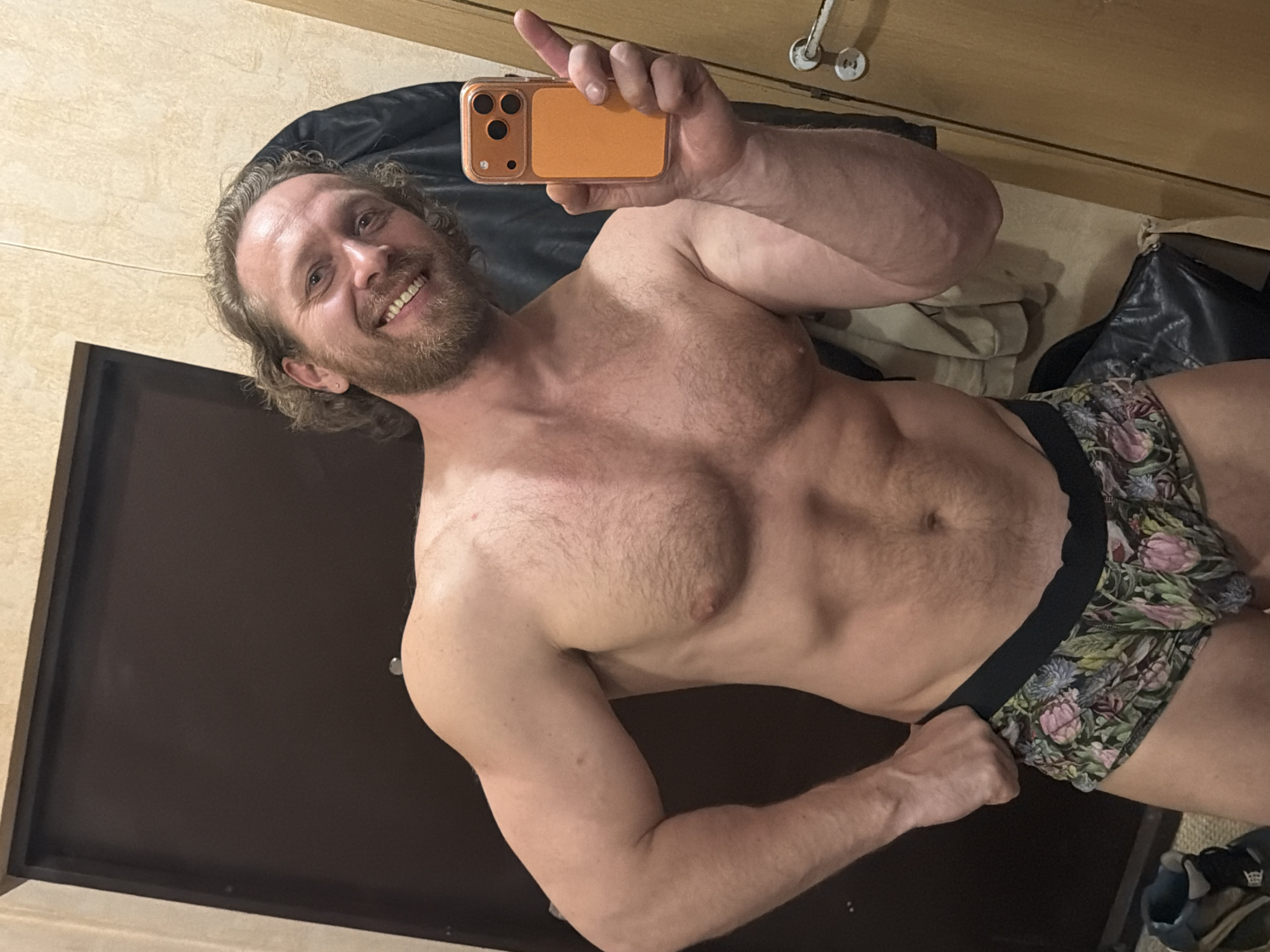 A shirtless man with a beard smiles into his phone, which he holds up in his right hand to take a selfie. He is wearing floral underwear and has a muscular physique. The background shows a room with a dark brown door, beige walls, and what appears to be a closet or storage area behind him with clothing and bags. The location cannot be reliably determined as Saint Petersburg, Russia based on this image alone.