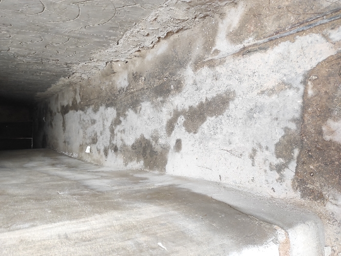 The image displays a narrow, concrete alleyway or corridor located within the city of Al-Qanawat Municipality, Syria. The scene is viewed from an elevated perspective, looking down the length of the passage into an unlit or dimly lit area, where a dark rectangular opening or door is barely discernible at the far end. The left wall is constructed from smooth, light gray concrete, exhibiting extensive dark, damp stains and streaks, particularly towards its base. The right wall features a dark gray, textured concrete surface with a repetitive, raised, wave-like or scale-like pattern. Its lower section is rough and appears partially eroded or unfinished. The concrete floor is a light gray, heavily marked with irregular dark, wet patches and accumulated dirt. A thin, dark linear object, resembling a metal pipe or wire, runs along the base of the right wall on the floor. A single white, small paper or plastic cup lies discarded on the floor towards the left side of the passage. There are no individuals, visible actions, or interactions present in the scene.