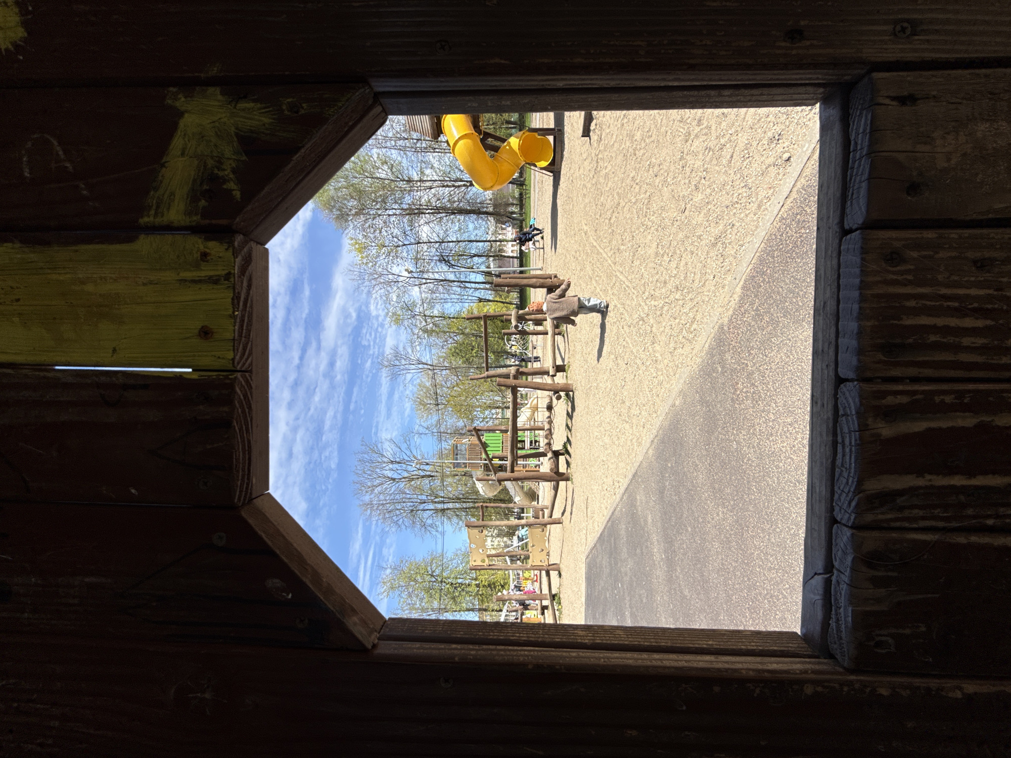 View from inside a wooden structure overlooking an outdoor playground in Заволжье, Russia. A yellow slide is visible in the background. A man in a jacket is lying prone, possibly on a piece of playground equipment. Another person is standing near the playground. The sky is blue with scattered clouds.