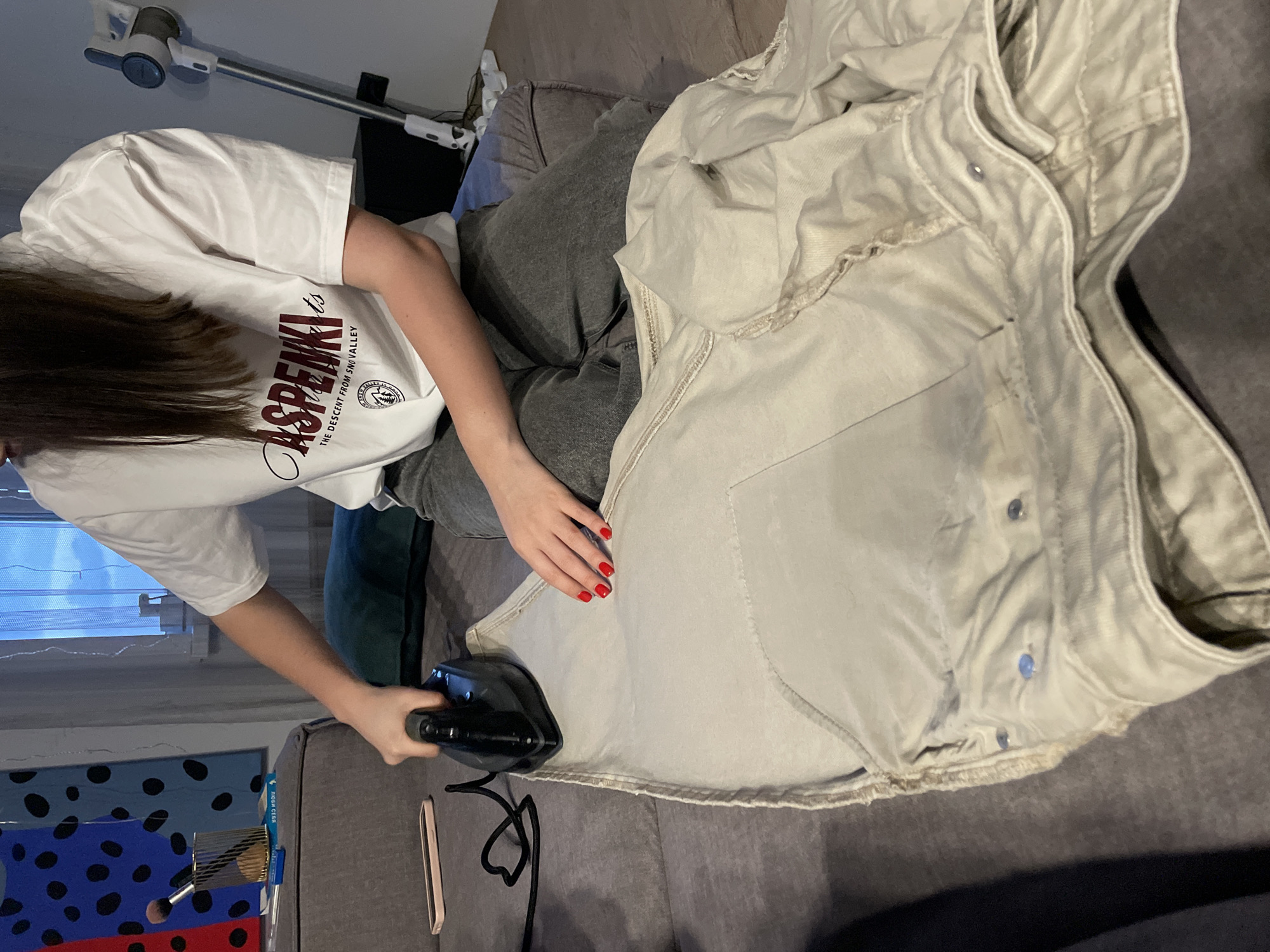 A person with long brown hair, wearing a white T-shirt with "Aspenki" written on it, grey sweatpants, and red nail polish, is ironing a light-colored garment, possibly a jacket, on a grey surface that looks like a sofa or bed. A black iron is in their right hand. A smartphone or case is on the surface near the iron. In the background, there is a window with a partially visible patterned piece of artwork or furniture and a portable vacuum cleaner. The scene is indoors, possibly a private residence. There is no explicit evidence to place the location in Krasnoyarsk, Russia.