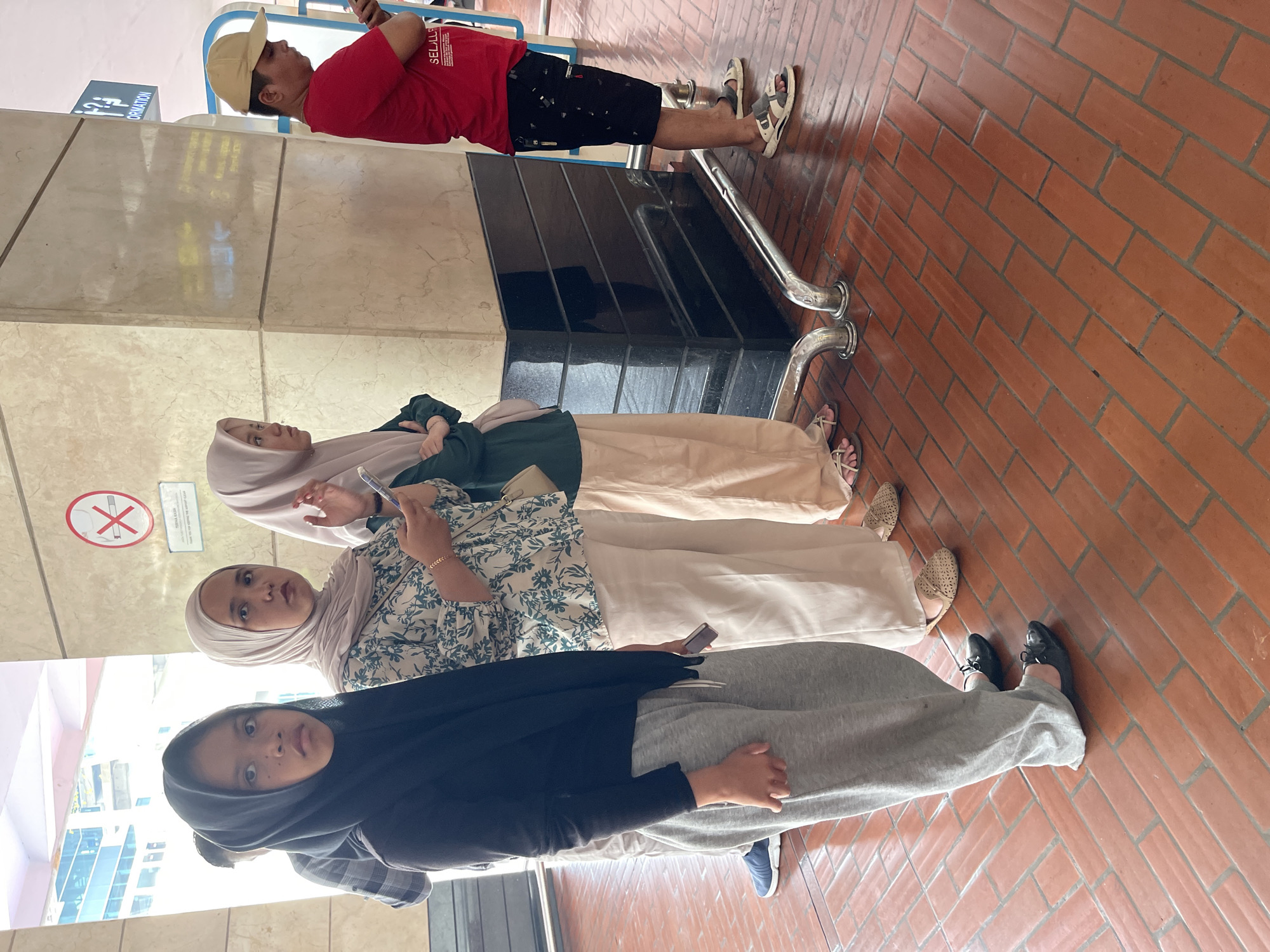 In Tangerang, Indonesia, a group of four people are standing in line on a reddish-brown tiled floor. The first person in line, from the left, is a young girl wearing a black hijab and grey sweatpants. The second person is a woman in a light-colored hijab and a floral patterned blouse, holding a smartphone. The third person is a woman wearing a light purple hijab and a green jacket, also with a handbag. The fourth person, on the far right, is a man wearing a red t-shirt, black patterned shorts, and a yellow baseball cap. He is looking at his phone. Behind the people, there is a large pillar with beige marble-like cladding. A silver railing runs along the floor in front of the pillar. Above the pillar, a sign with an information symbol and text is visible. Another sign with a red circle and a diagonal line through a cigarette symbol indicates a no-smoking area. The environment appears to be an indoor public space, possibly a transportation hub or a shopping mall.