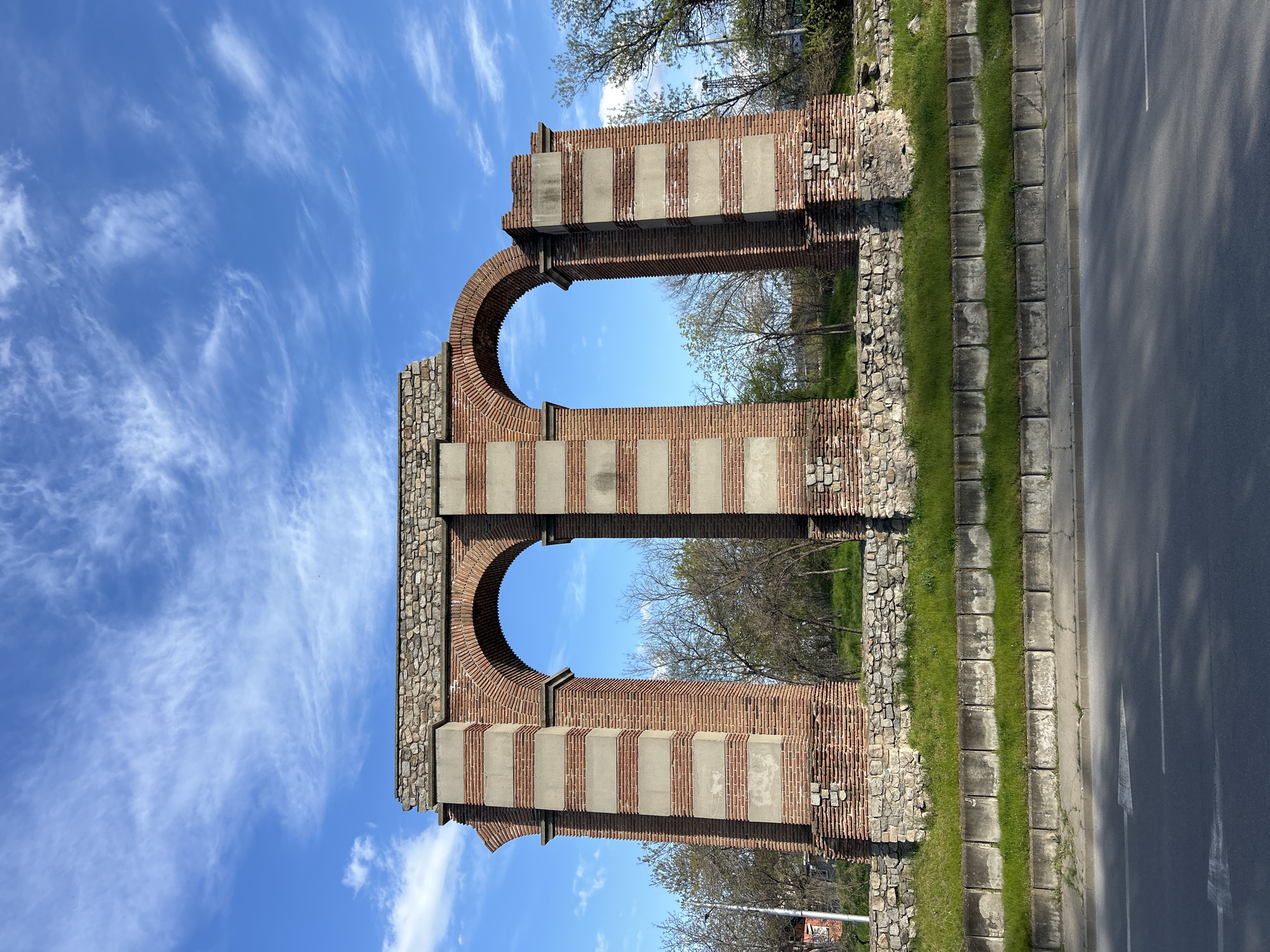 The image shows the remains of the Roman aqueduct in Plovdiv, Bulgaria. The structure is made of brick and stone, with two arched openings visible against a blue sky with cirrus clouds. The aqueduct is situated next to a road, separated by a strip of grass and a curb. Some trees are visible behind the structure.