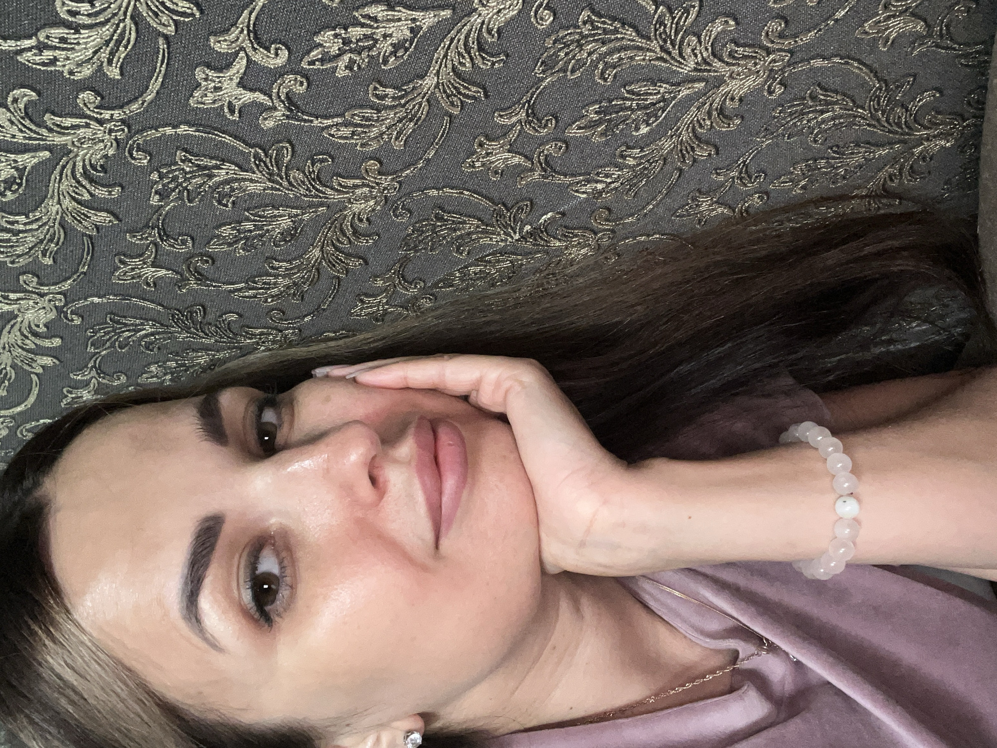 A woman with long, brown hair is pictured from the chest up, resting her chin on her hand. She is wearing a pink t-shirt, a delicate necklace, and a bracelet made of light-colored beads. A diamond stud earring is visible in her left ear. The background is a dark, textured wallpaper with an ornate, gold floral pattern. The woman's face is turned slightly towards the camera, with her eyes looking forward. The lighting is soft, highlighting her features and the details of the wallpaper.