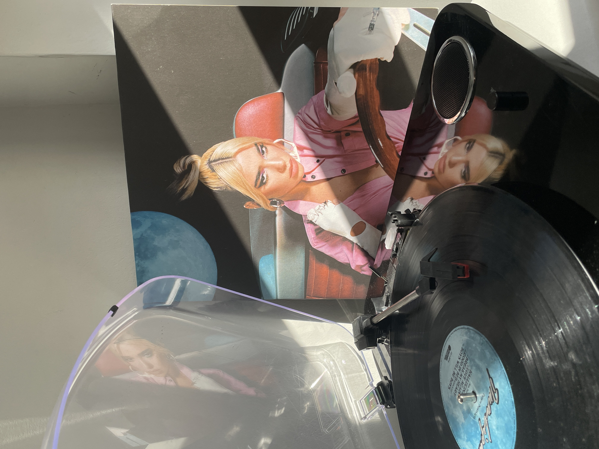 Here's a detailed description of the image:
The image shows a vinyl record player playing a record of Dua Lipa's "Future Nostalgia" album. The album cover is visible behind the record player. The record player is black and has a clear plastic cover. The vinyl record itself is black with a blue label. The label includes the album title and a list of songs including: "Hallucina", "Break My Heart", "Love Again", "Boys Will Be Boys", and "Good In Bed". The cover of the "Future Nostalgia" album features an image of Dua Lipa wearing a pink outfit and white gloves while seemingly sitting in a car. A blue moon is visible in the background.
The scene is indoors and well-lit.