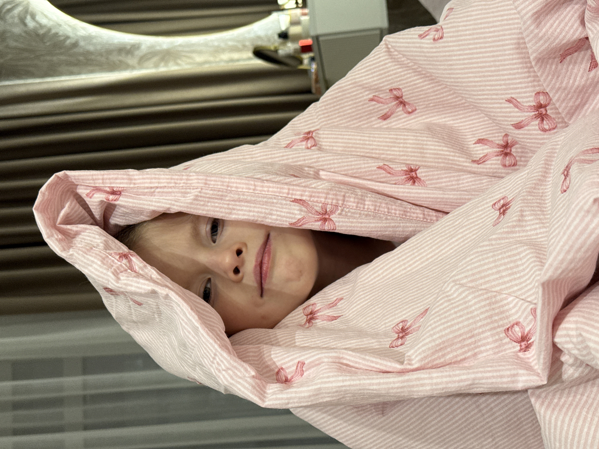 A young child lies under a pink and white striped blanket patterned with pink bows. The child's face is visible, and they appear to be looking directly at the viewer. The setting is indoors, possibly a home. The location is unknown, and there is no evidence the photo was taken in Izhevsk, Russia.