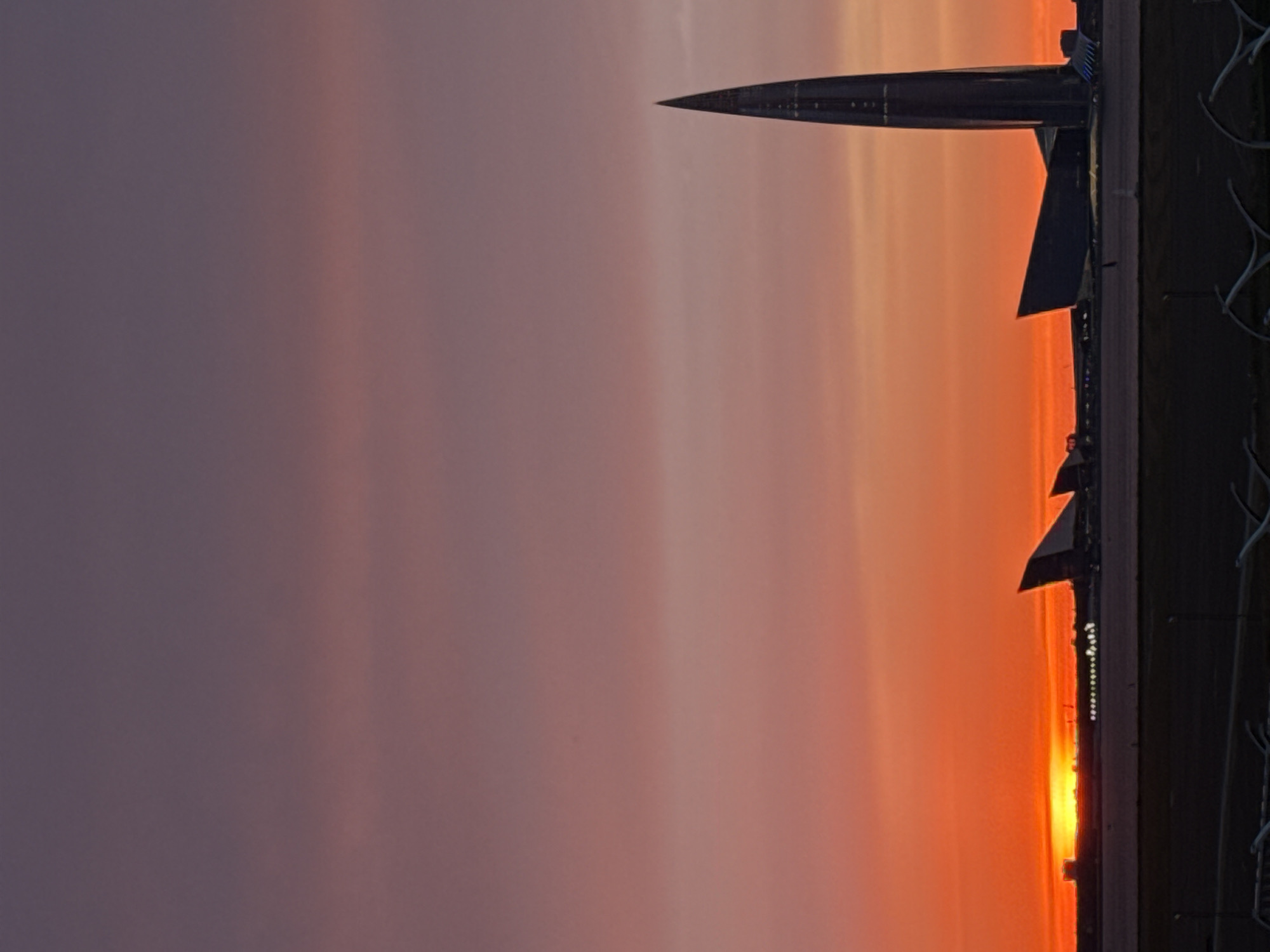 This vertical, wide-angle photograph captures a sunset over a city skyline, with a prominent modern skyscraper and other architectural structures silhouetted against a gradient of orange, pink, and purple hues in the sky. The sunset is vivid, with the sun appearing as a bright orange band low on the horizon, casting an intense glow. To the right of the frame, a tall, slender, pointed skyscraper, resembling a shard or needle, dominates the view. This is the Lakhta Center, a landmark in Saint Petersburg, Russia, located in the northwestern part of the city on the Gulf of Finland. To the left of the skyscraper, several other angular, modern buildings are visible, also in silhouette. The foreground of the image shows a dark, flat expanse, possibly an airport or an open waterfront area, with a few low-lying structures and some indistinct fencing or barriers. The overall atmosphere is dramatic and serene, characterized by the striking colors of the sky and the stark silhouettes of the urban architecture.