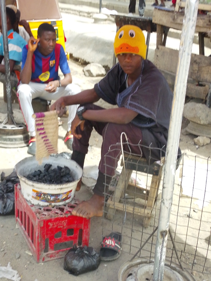 The image depicts an outdoor scene in Fagge C, Nigeria. In the foreground, a male individual with dark skin sits on a low wooden stool. He is wearing a yellow knitted beanie with a duck face design, a dark short-sleeved garment with blue trim, and dark trousers. His right hand holds a small, woven, rectangular mat-like object with purple patterns. He is barefoot, and a single black slide sandal with "ABA" branding rests on the ground next to his left foot. Immediately in front of him, a white plastic bucket filled with black charcoal is placed on top of a red plastic crate. A small black plastic bag lies on the ground beside the crate. A metal grate structure is positioned directly in front of the seated individual. In the midground, to the left, a second male individual with dark skin is seated on an upturned metallic wheel rim. He is wearing a blue and red striped short-sleeved shirt and white pants with red side stripes. His left hand is raised near his ear. The background includes indistinct figures, simple wooden structures, and a partial view of a yellow vehicle on the far left. The ground is an unpaved or lightly paved surface.