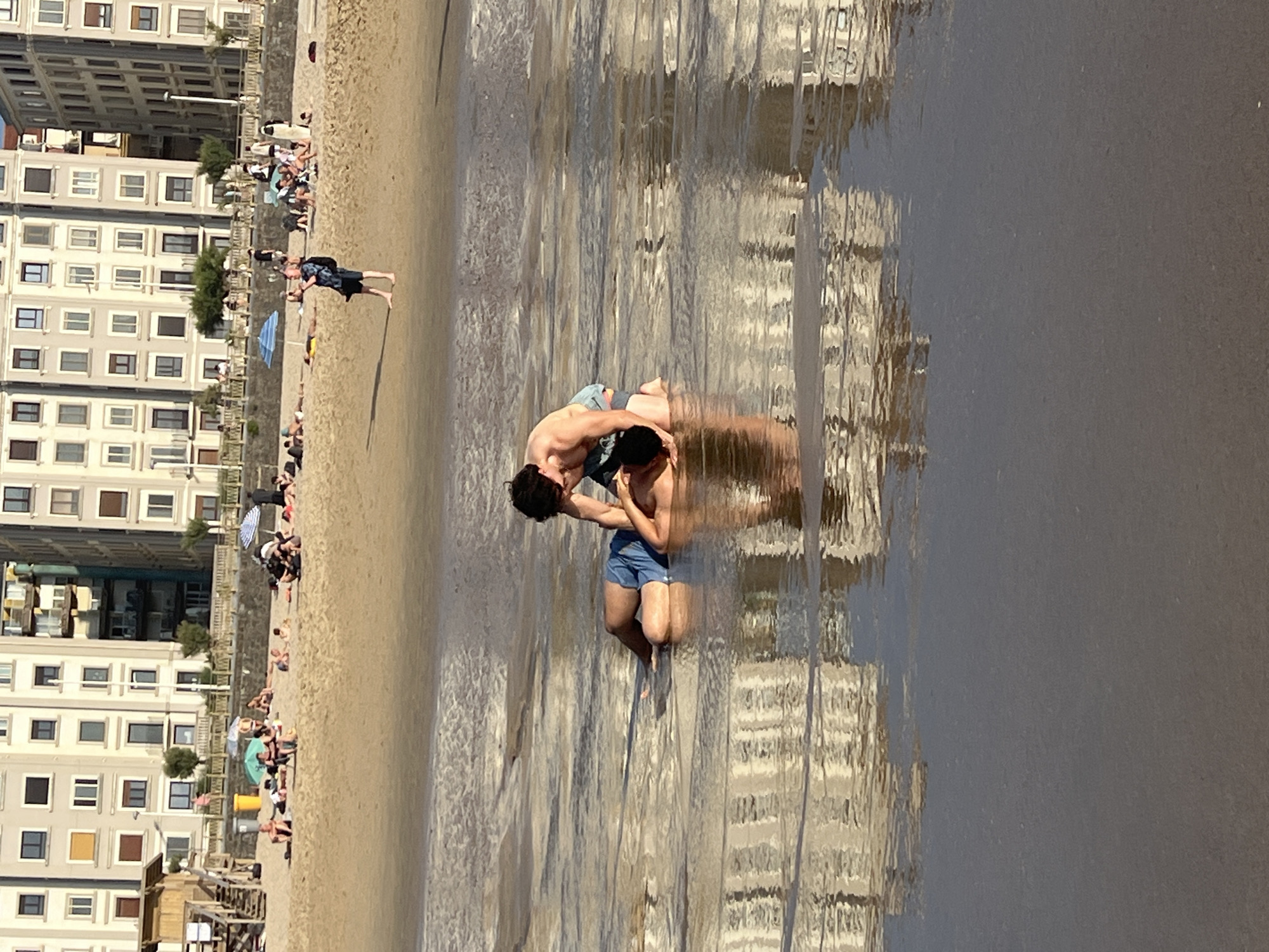 Two men are in shallow water on a beach, with one man kneeling and the other lying on his back. The kneeling man appears to be helping the lying man. The beach is sandy, with buildings in the background that suggest an urban environment. The water reflects the buildings. In the background, other people are visible on the beach, some are sitting or lying down, and others are walking. Beach umbrellas and a small lifeguard tower are also present. The scene suggests a warm day at a beach. The architecture of the buildings suggests a European city, and the presence of a beach is characteristic of coastal cities. Given the context of San Sebastián, Spain, this scene is likely La Concha beach, a well-known urban beach with a prominent promenade and buildings.