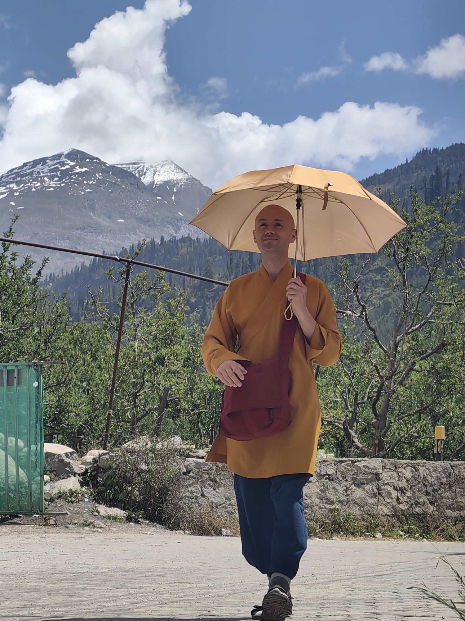 A full-body, medium shot shows a bald man wearing a saffron robe and dark blue pants, walking on a dirt path with a light brown umbrella open above him. He is carrying a maroon bag slung across his body. The man is looking up and to his left with a slight smile. Behind him, a rocky wall with sparse vegetation borders a slope covered in trees. In the far background, a snow-capped mountain rises under a blue sky with scattered white clouds. A green metal fence is visible to the left of the man. The scene appears to be outdoors in a mountainous region, possibly in or near Barheta, India, given the landscape.