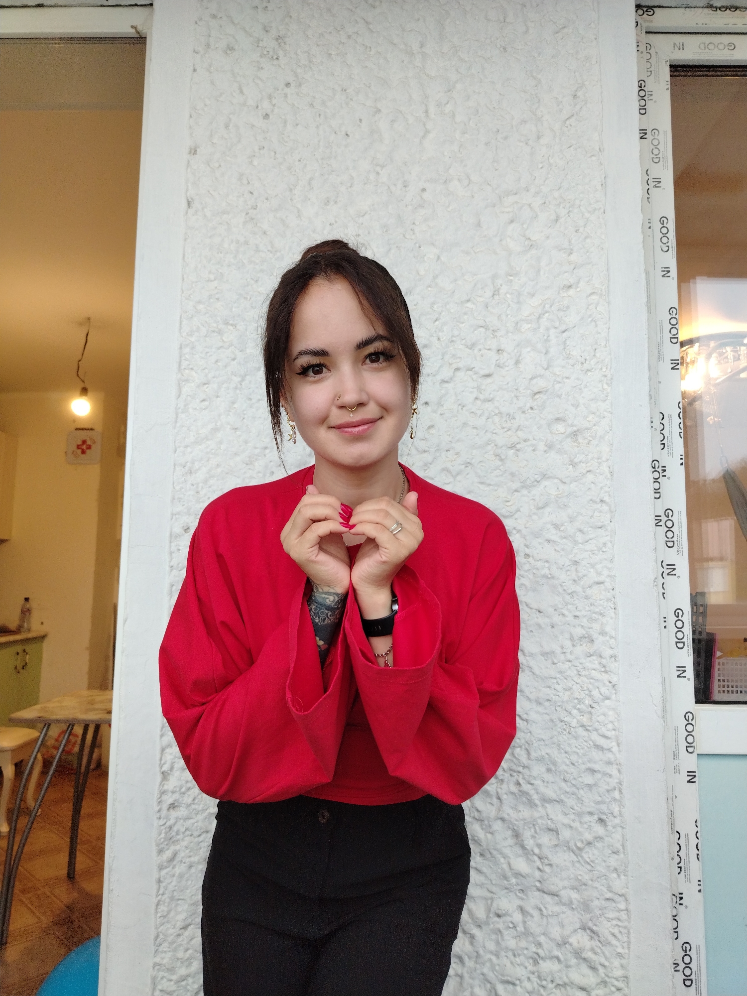 A young woman with dark hair pulled back in a bun is standing in front of a textured white wall, possibly a building exterior. She is wearing a red long-sleeved shirt and black pants. Her hands are clasped together in front of her chest, forming a heart shape. She has a nose ring and delicate earrings. To her left, a doorway or window reveals an interior space with furniture and a light fixture. To her right, a window frame with repeated text and graphics is visible. The background suggests an urban or semi-urban setting.