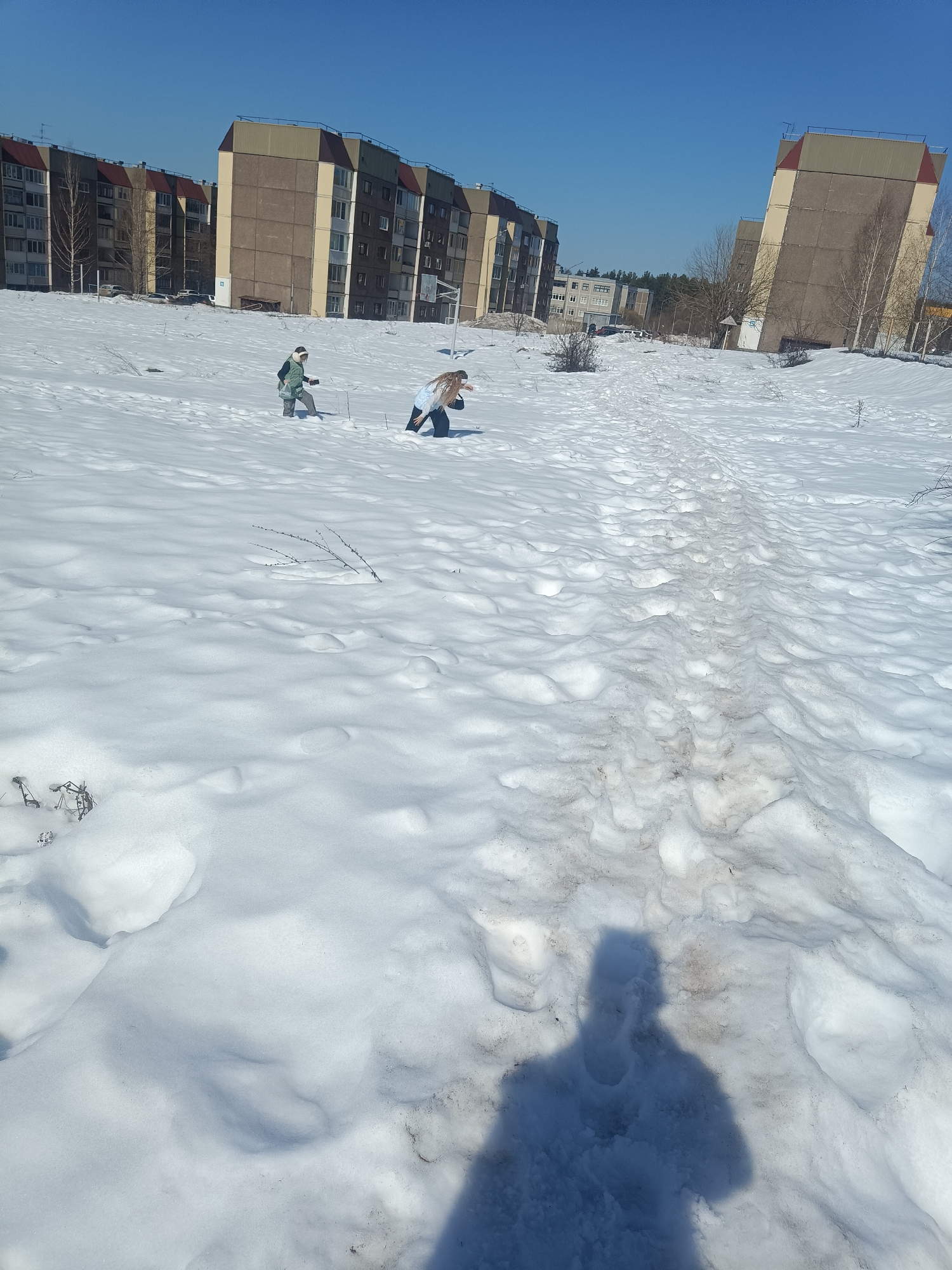 The image shows a snowy landscape with several buildings in the background, likely in the city of Марковский, Russia. Two individuals are present in the mid-ground. One individual, wearing what appears to be a green coat or vest, is standing. The second individual is in a kneeling position. A path worn through the snow is visible, leading from the background towards the foreground. The shadow of the photographer is visible in the foreground. The sky is clear and blue. The buildings are multi-story and appear to be residential or commercial structures. Vegetation is sparse, with some bare trees and shrubs visible.