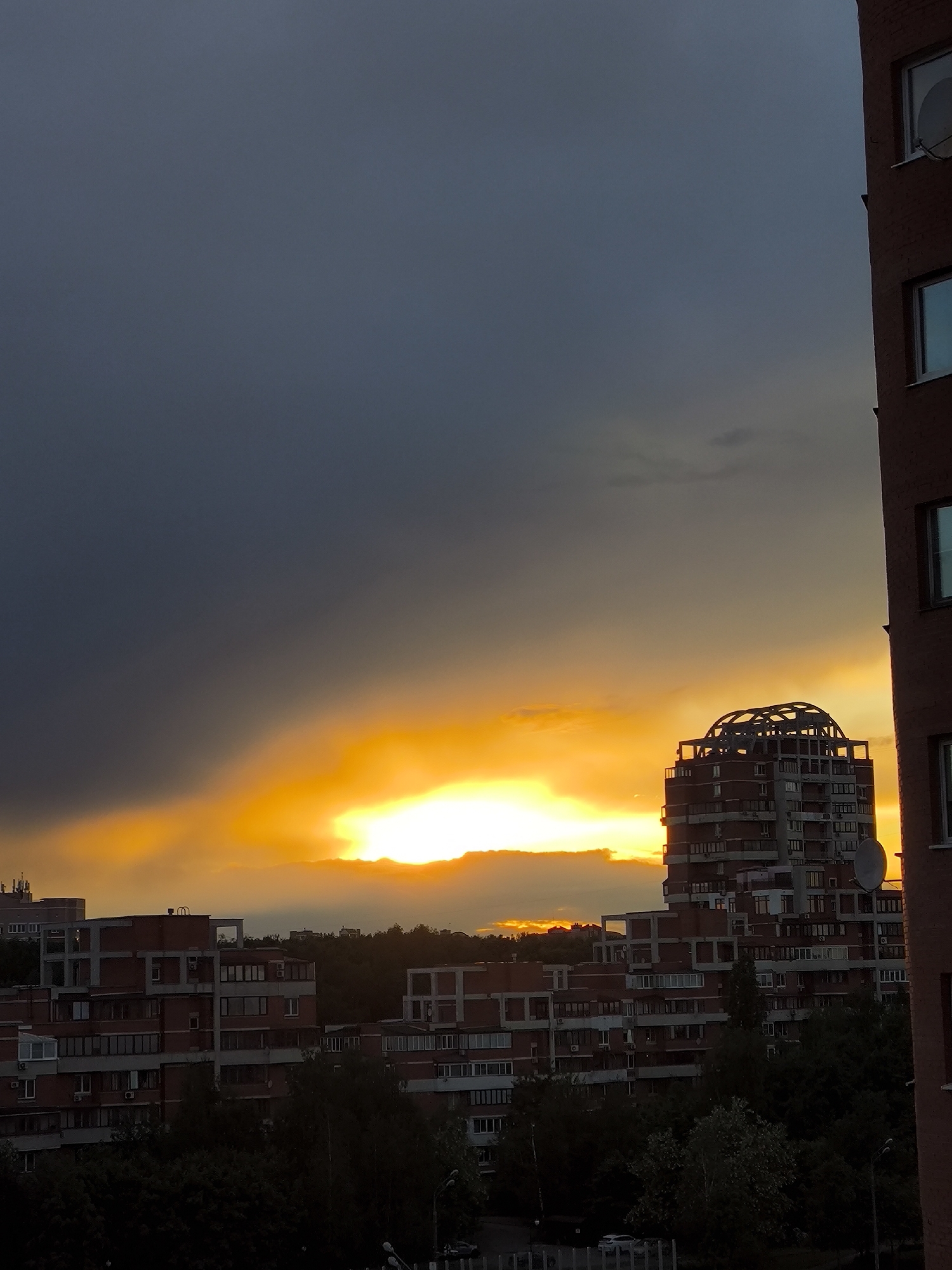 The image depicts an urban scene at sunset in an unknown city. Dark, stormy clouds fill the upper portion of the frame, contrasting sharply with a vibrant orange and yellow sky where the sun is setting. In the foreground, several multi-story brick buildings with numerous windows are visible. The tallest building is a unique structure with a dome-like unfinished top, possibly under construction. Trees are scattered around the base of the buildings. A corner of another building is visible on the right edge of the image.