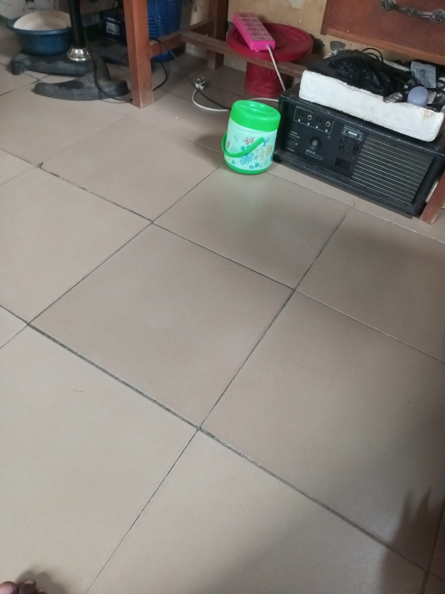 An indoor scene features a floor tiled with large, square, light brown tiles. In the background, a wooden table leg is visible, supporting a wooden surface. To the left, a black fan with a wide base stands on the floor. On the table, a blue bowl containing white powder and a blue basket are present. To the right of the fan, on the floor, sits a green plastic bucket with a white lid and a handle. Further to the right, a black amplifier is placed on the floor, topped with a white foam block and a black object. A pink remote control sits on a red disc on the table. The scene appears to be in Ihalav, Nigeria.