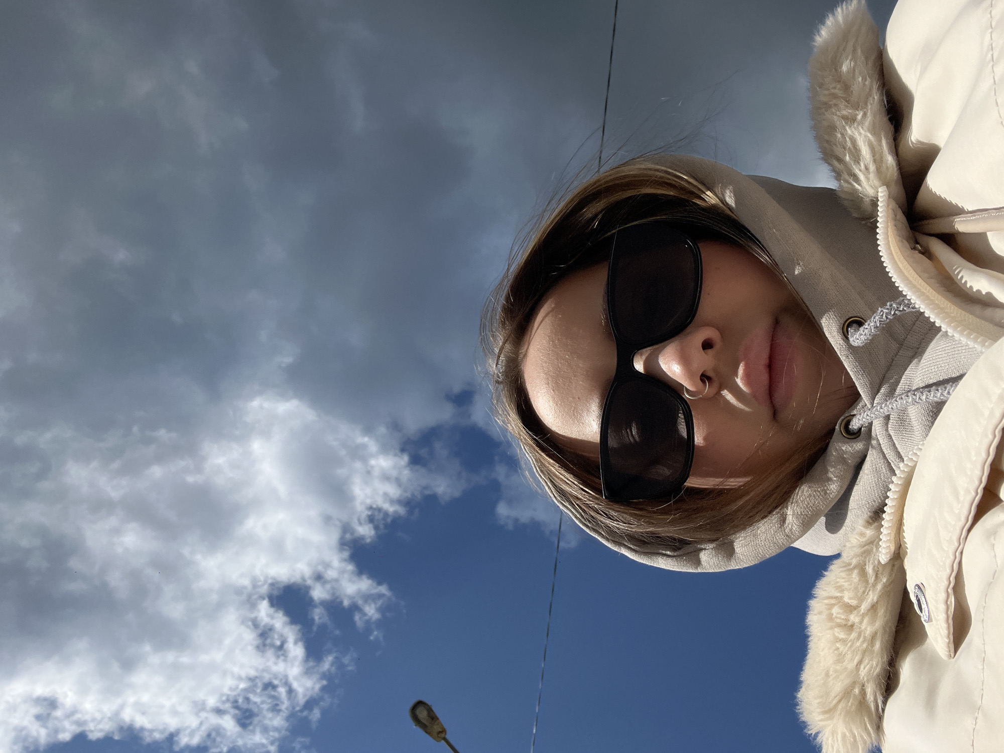 A low-angle shot captures a person wearing dark sunglasses and a hooded jacket with a fur trim. The background features a partly cloudy sky transitioning from gray to blue. A street lamp post and power lines are also visible. The image was possibly taken in Novopokrovskaya, Russia.
