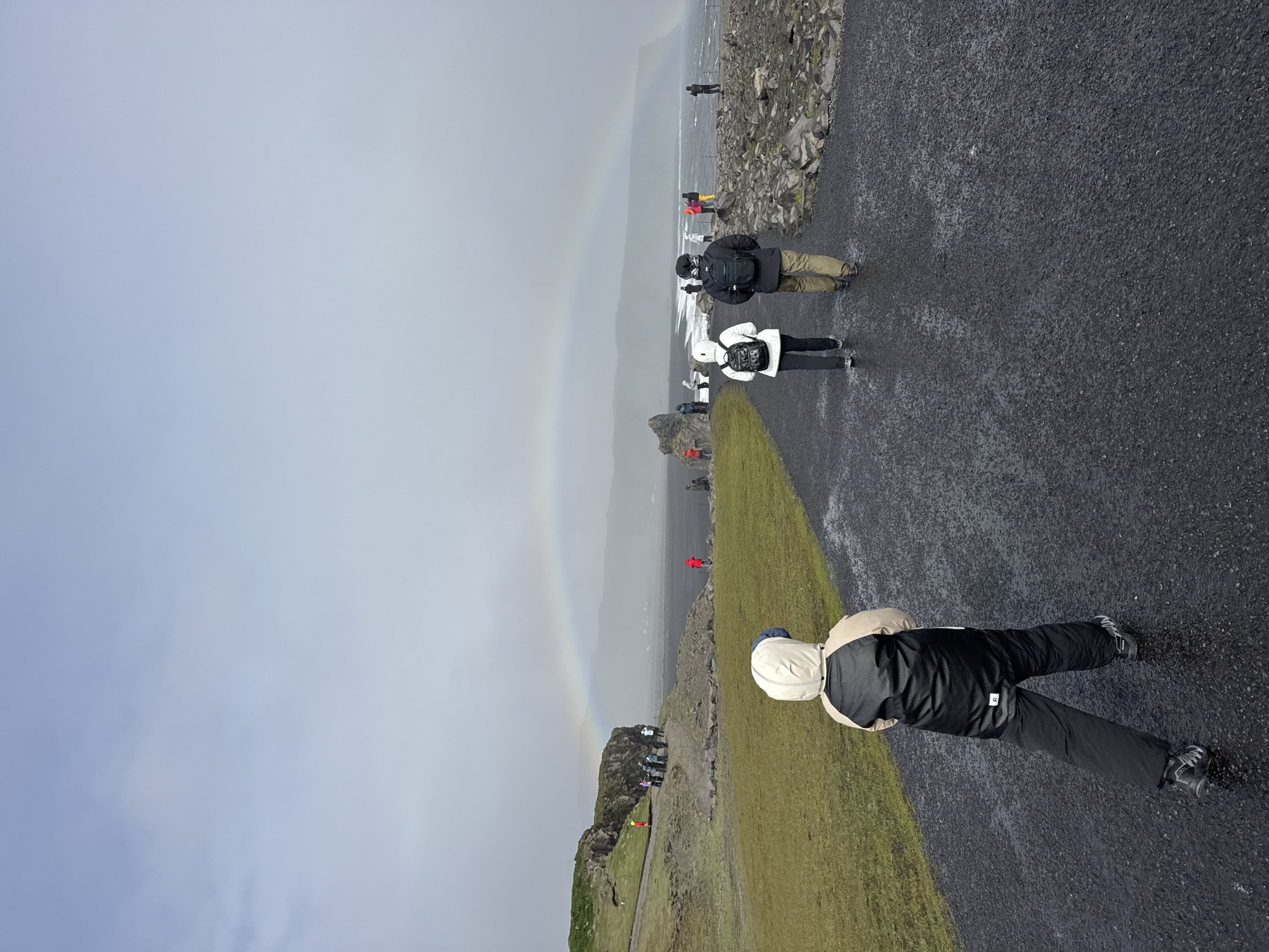The image depicts a landscape scene in Vik, Iceland, featuring a rainbow extending from the sky to the ground amidst a grey and overcast day. A group of people, tourists perhaps, are positioned along a black paved path that borders a grassy area. One person is close to the camera with their back to the viewer, wearing a light-colored jacket and dark pants. Further away, other individuals can be seen walking along the path and some standing near a rocky cliff overlooking a black sand beach and ocean. The landscape is marked by volcanic rock formations. The overcast sky obscures sunlight and emphasizes the colors of the rainbow.