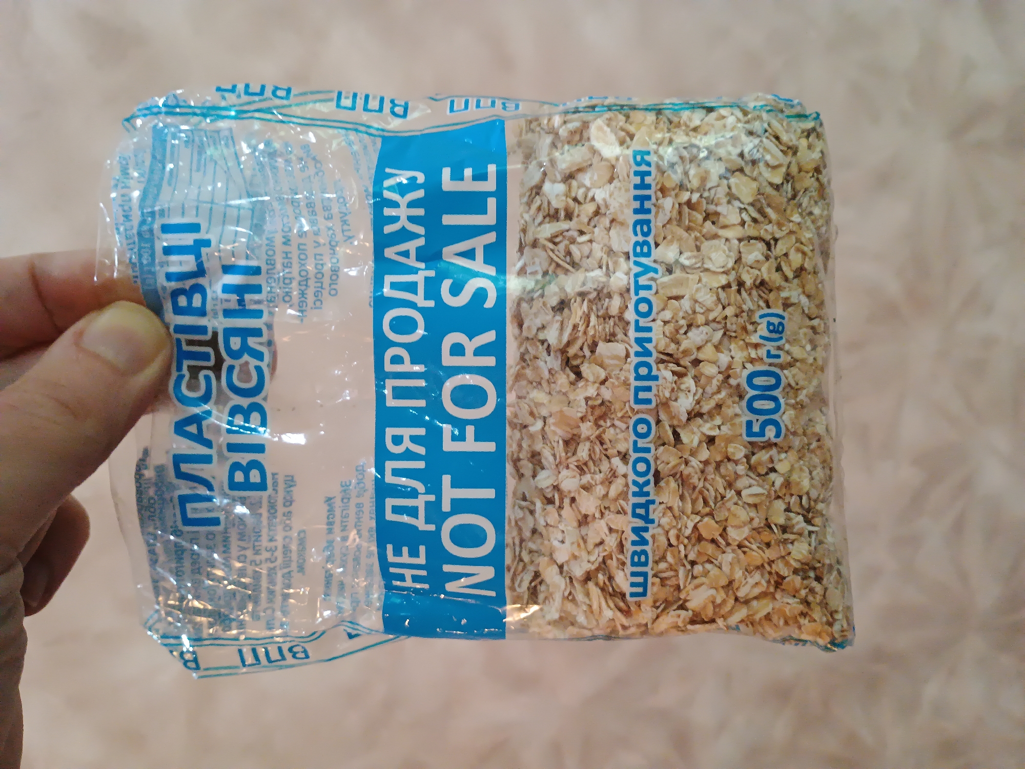 The image shows a bag of oatmeal held by a hand. The bag has text in Ukrainian and English. The Ukrainian text reads "ПЛАСТІВЦІ ВІВСЯНІ" (oat flakes) and "НЕ ДЛЯ ПРОДАЖУ" (not for sale). The English text reads "NOT FOR SALE". Below this, in Ukrainian, it says "швидкого приготування" (quick cooking). The bag also indicates a weight of "500 г (g)". The background is a light-colored wall. The image does not provide any information about the city of Zaporizhzhia, Ukraine.