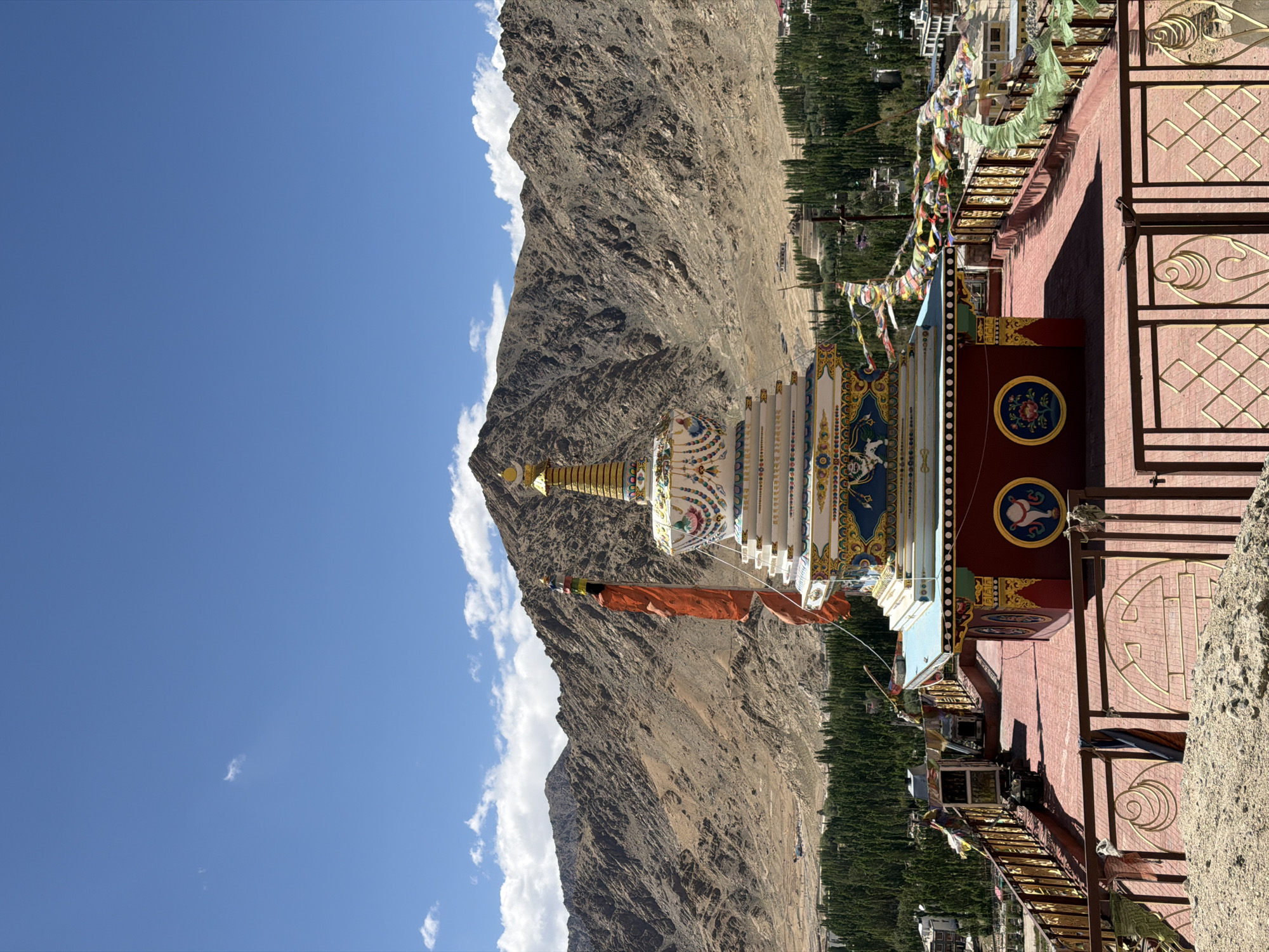 A stupa, adorned with colorful prayer flags, stands on a rooftop overlooking the town of Leh, India. The stupa is white with gold accents, intricate blue and red detailing, and a golden spire topped with a crescent moon. An orange flag flutters from a pole beside the stupa. The rooftop is tiled in red and surrounded by a decorative metal railing. In the background, a range of barren, rocky mountains rises against a bright blue sky dotted with white clouds. A lush green belt of trees separates the town from the mountains, with buildings visible among the trees.