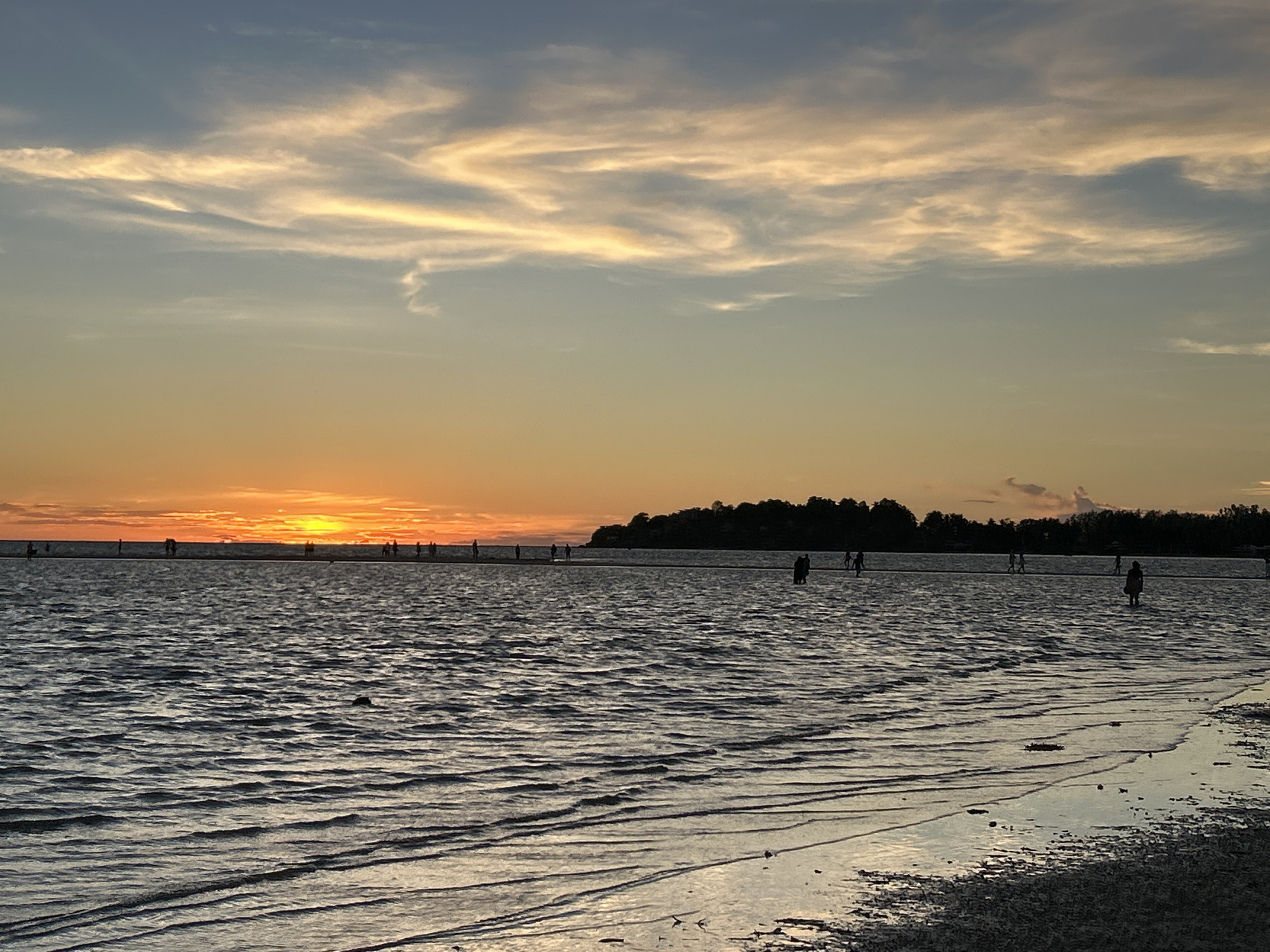 This image depicts a serene beach scene at sunset in Koh Phangan, Thailand. The sky is painted with hues of orange, yellow, and blue, with wispy clouds illuminated by the setting sun. The sun itself is a bright orange orb low on the horizon, casting a warm glow over the water.
The foreground is dominated by the calm, rippling surface of the sea, reflecting the colors of the sky. The shallow water extends towards a distant sandy bank or sandbar where several people are visible, silhouetted against the bright sunset. They appear to be leisurely walking or standing in the water, enjoying the twilight ambiance.
Further in the distance, a line of trees forms a dark silhouette against the horizon, suggesting a landmass or island. The overall atmosphere is tranquil and peaceful, evoking a sense of calm and relaxation often associated with beach sunsets. The weather appears clear and pleasant, ideal for an evening stroll by the sea. There is no visible text in the image.