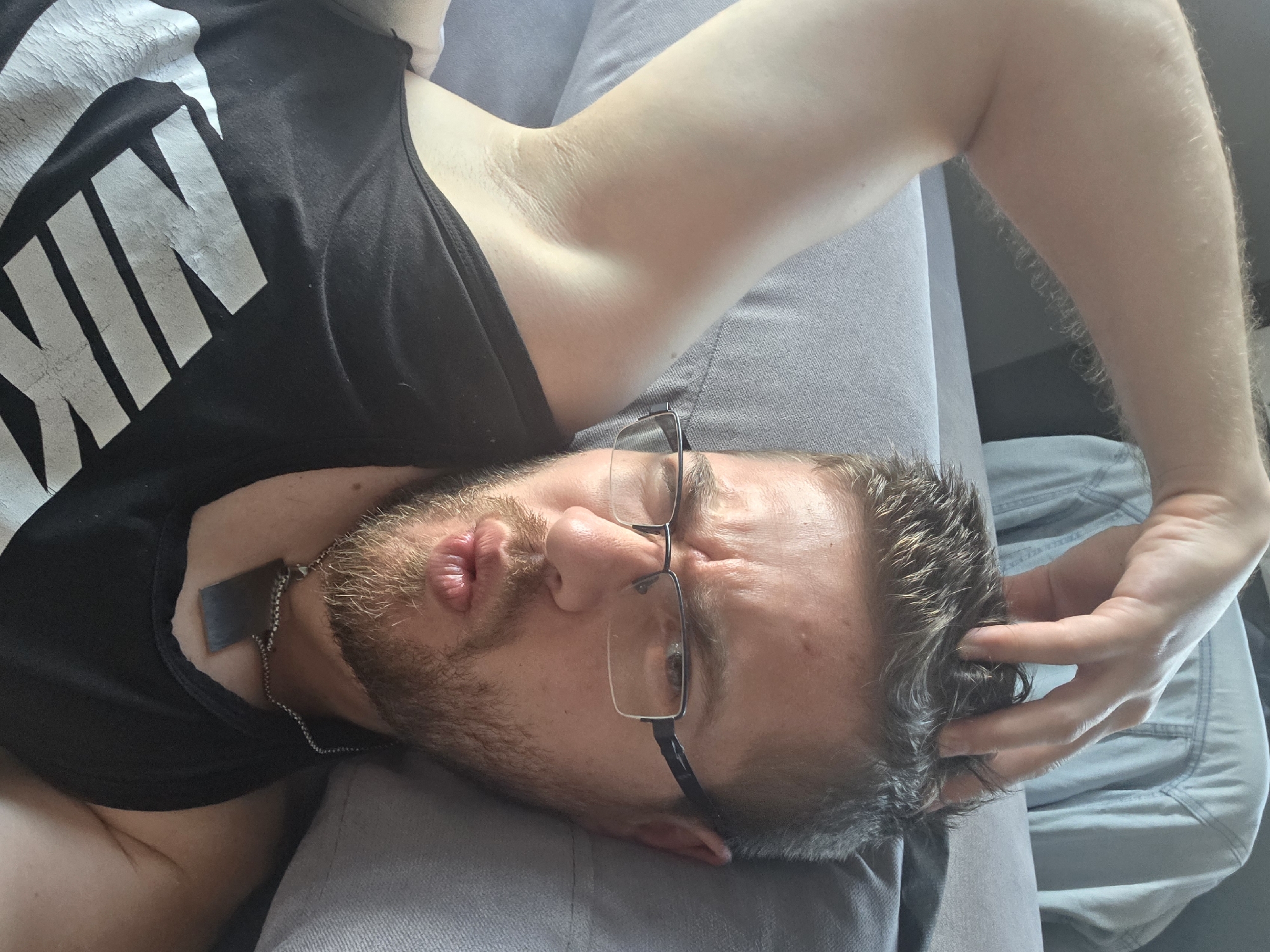 An adult male with light skin, short brown hair, and a short beard and mustache lies on a light gray upholstered surface, viewed from a high angle. He wears rectangular-framed glasses with dark rims and a silver bridge. His right arm is bent with his hand on top of his head, fingers resting in his hair. His lips are pursed in a "fish face" expression, and his eyebrows are slightly raised, creating minor furrows on his forehead. He wears a black sleeveless tank top with a faded white "NIKE" logo on the chest, and a silver-colored chain necklace with a flat, rectangular, dark metallic pendant around his neck. In the upper background, a light blue denim shirt is draped over an unseen object. The scene takes place indoors in Yagodovo, Bulgaria.