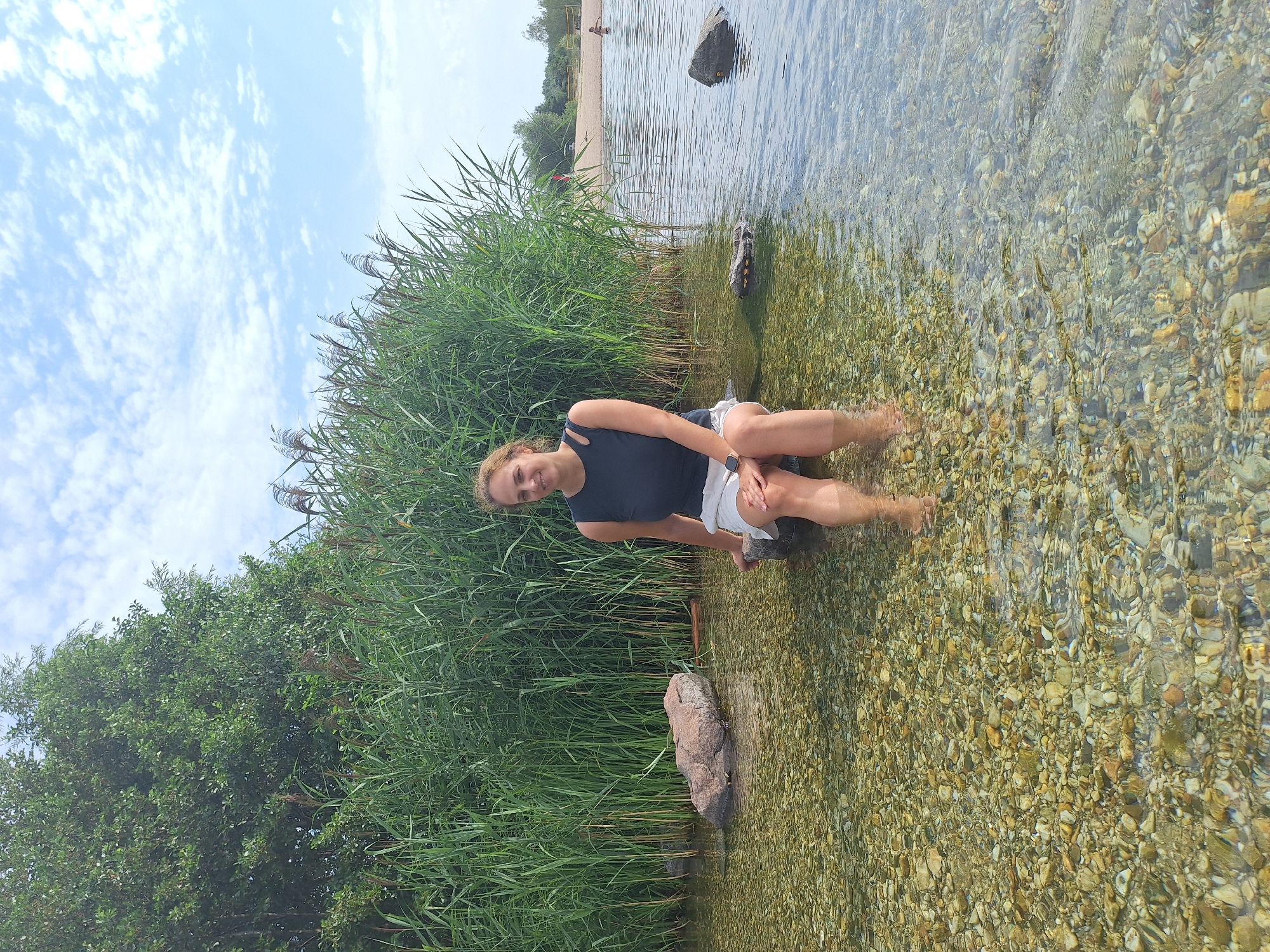 A woman is sitting on a rock in shallow water with reeds behind her and a sandy beach in the distance. She has blonde hair and is wearing a black top and white shorts. The water is clear and shows the rocky bottom. The sky is blue with white clouds. The location is likely a lake or riverfront area, possibly within or near the city of Leipzig, Germany, given the presence of reeds and a beach.