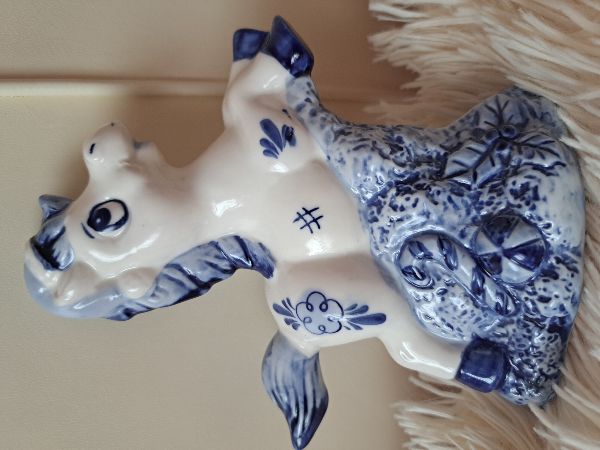 A close-up, eye-level, medium shot captures a white ceramic figurine of a horse with blue decorative details. The figurine is adorned with intricate blue patterns, including floral motifs, a crosshatch symbol, and splashes of color on its mane, tail, legs, and hooves. The horse is positioned on a textured, blue and white base that resembles rocky terrain, with depictions of leaves and circular patterns. The figurine rests on a fluffy, cream-colored surface, likely a rug or blanket, which extends into the foreground and background. The background is a plain, textured surface, possibly a wall or furniture. The lighting appears soft and diffused, casting subtle shadows. There are no people or animals present in the image. This scene is not located in the city of Irkutsk, Russia.