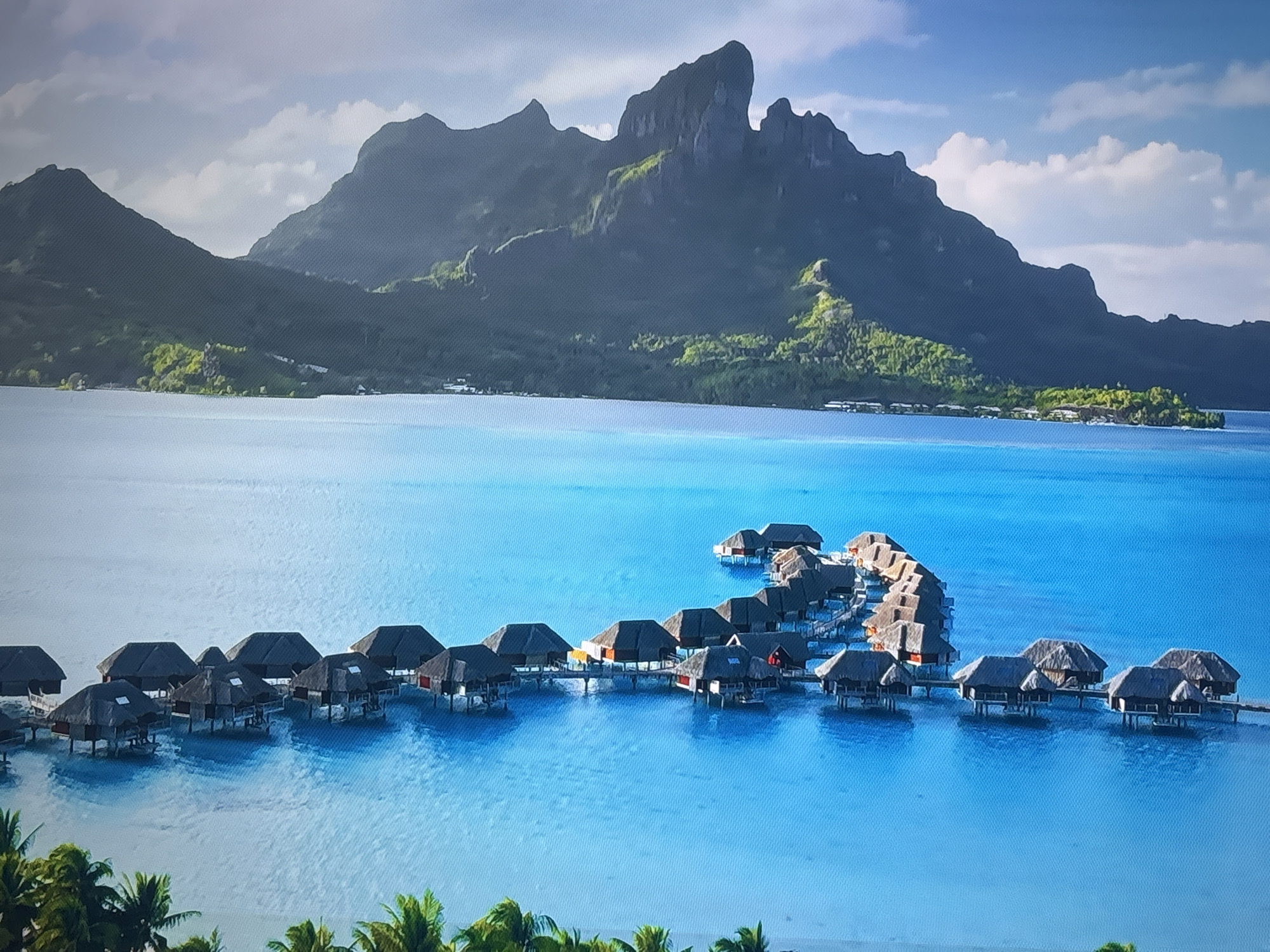 This media file depicts an aerial view of an overwater bungalow resort situated in a clear, turquoise lagoon. The scene is dominated by the vibrant blue water and the thatched-roof bungalows, many of which are connected by wooden walkways. The bungalows are clustered together, forming a unique architectural pattern on the water. In the background, a lush, mountainous island rises from the sea, its peaks shrouded in soft clouds under a bright, partly cloudy sky. The greenery on the island contrasts with the deep blue of the ocean and the muted tones of the mountains. The time of day appears to be morning or early afternoon, with the sun casting a gentle light on the water and landscape. There are no visible people or text in the image, and the overall mood is one of tranquility and tropical paradise. The setting is a serene coastal environment, likely a tropical island known for its scenic beauty and luxury resorts.