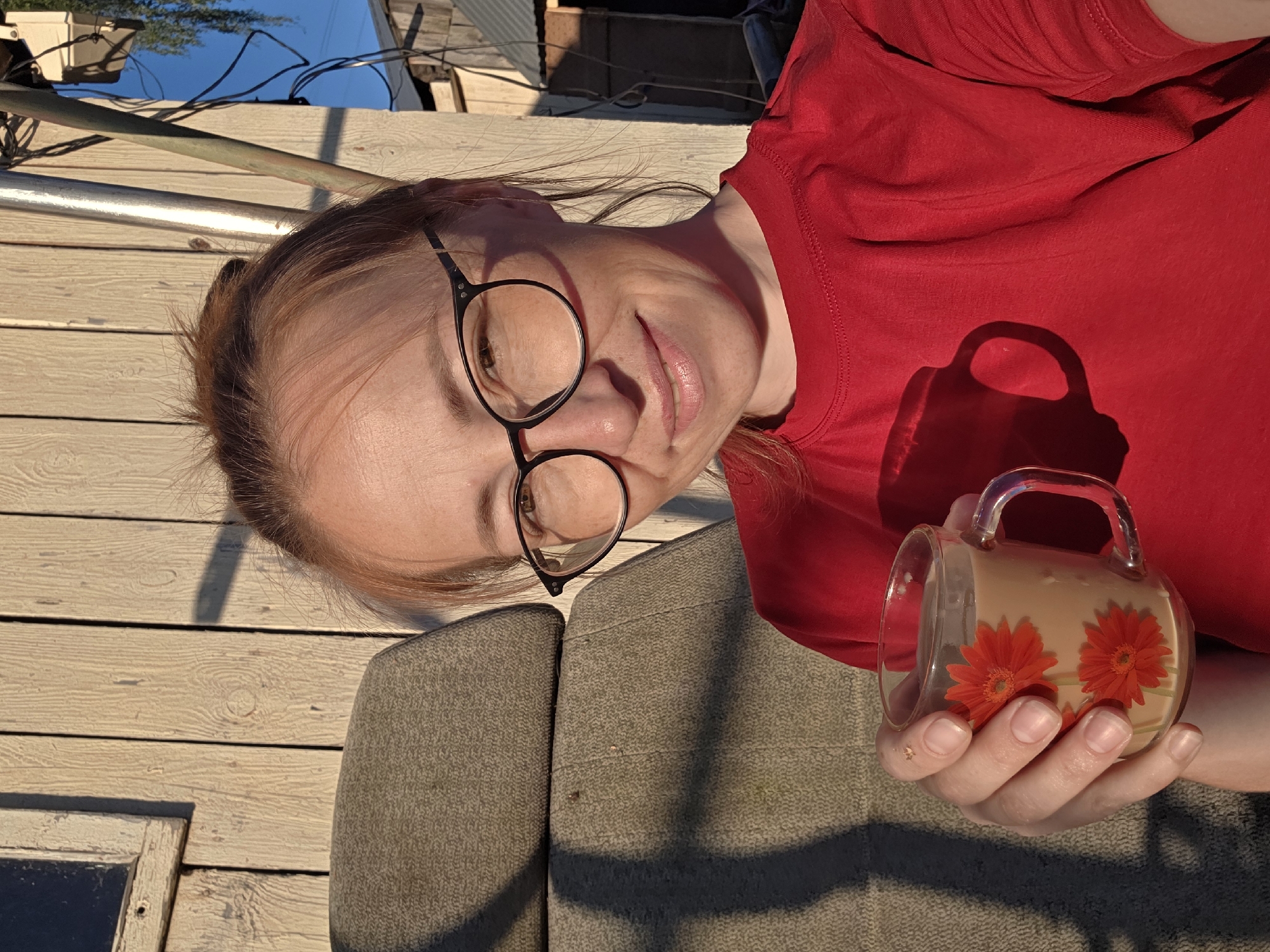 This is a close-up, rotated image of a woman lying down, holding a mug. She has light skin, reddish-brown hair, and is wearing black-rimmed glasses and a red t-shirt. Her eyes are looking towards the camera, and she has a slight smile. The mug is clear glass with a handle and contains a light brown liquid. Two red flowers are depicted on the exterior of the mug.
The setting appears to be outdoors, with wooden planks forming a surface beneath and behind the woman. Sunlight casts shadows, suggesting it is either morning or late afternoon. In the background, there are glimpses of what looks like a building with a corrugated roof and wires, along with a blue sky. The overall mood is relaxed and casual. The location context provided is "Буренка, Russia." There is no visible text in the image.
