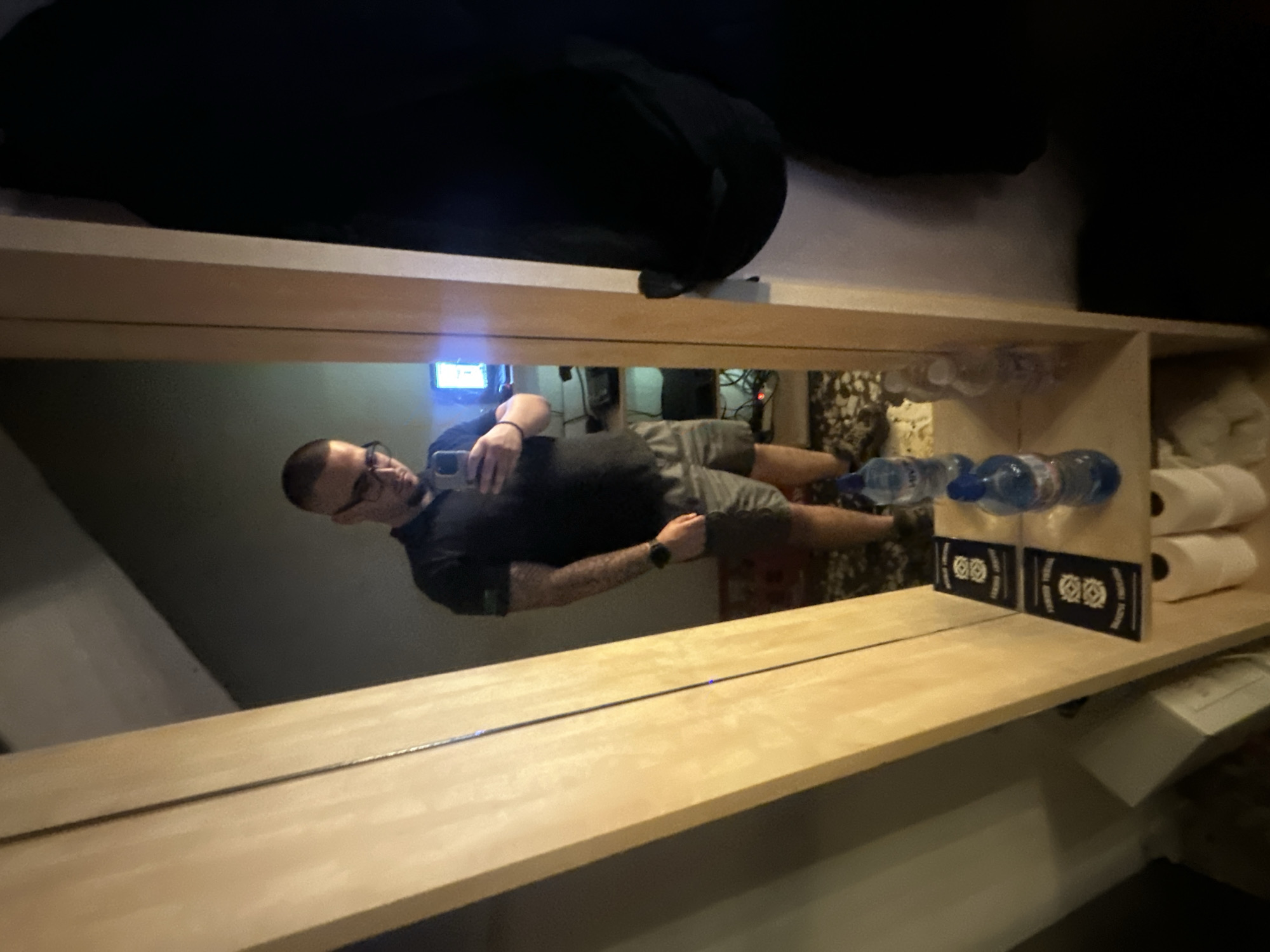The image captures a mirror selfie taken by a man in an indoor setting, likely a hotel room in Lom, Bulgaria. The man, reflected in a large rectangular mirror, has short hair, a beard or stubble, and wears glasses. He is dressed in a dark short-sleeved t-shirt and grey shorts. Tattoos are visible on his left arm, and he wears a watch on his left wrist. He is holding a smartphone in his right hand, positioned to take the photograph.
The reflection shows a light-colored wooden shelf or countertop running horizontally across the lower part of the mirror. On this shelf, several plastic bottles of water with blue caps are arranged. To their right, two rolls of white toilet paper are neatly stacked within a cubby or on a smaller shelf. Between the water bottles and the toilet paper, two small, dark-colored cards or signs featuring white circular designs are displayed.
In the background of the reflection, a glowing blue light from an electronic device is visible to the left of the man's head. Other details like cables and patterned curtains or fabric can be vaguely discerned, suggesting a furnished room. The overall dim lighting indicates an indoor setting, possibly evening or night. No specific text on labels or cards is clearly legible.