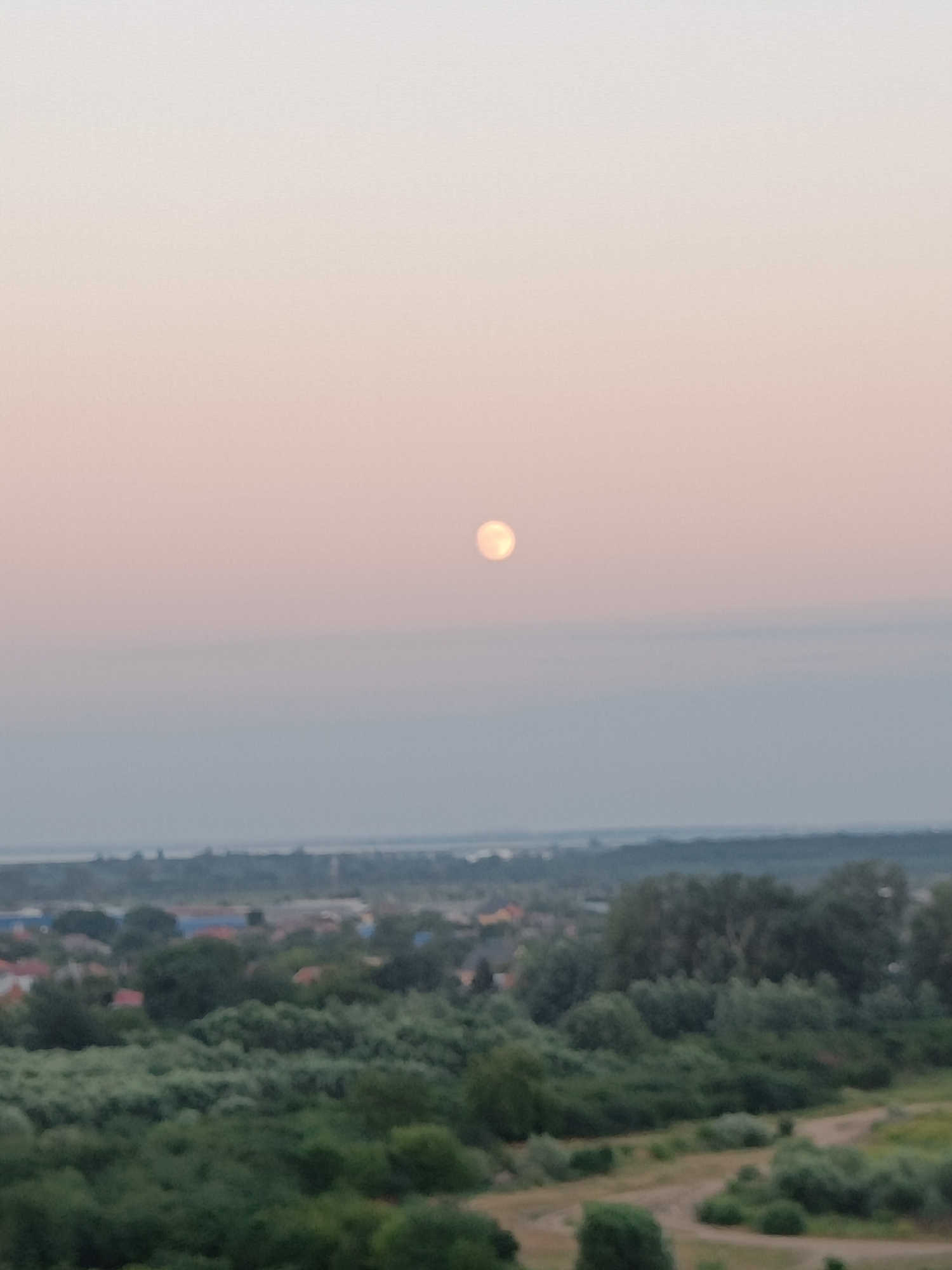This image captures a hazy, tranquil scene overlooking a landscape at dusk. The most prominent subject is a pale, glowing orb, likely the moon or a very low sun, positioned in the upper center of the frame against a soft, gradient sky that transitions from a pale pink at the top to a light gray towards the horizon.
Below the sky, a muted, distant view of a settlement is visible, characterized by rooftops and structures partially obscured by atmospheric haze. This settlement is bordered by a dense band of dark green trees, suggesting a rural or semi-urban environment. Further in the foreground, a foreground of lush green foliage and some bare earth with winding paths is depicted, indicating a natural or park-like area.
The overall atmosphere is serene and still, with the soft lighting and muted colors suggesting either dawn or dusk. Given the location context of Krasnodar, Russia, this could be a view from an elevated point looking out over the outskirts of the city or a nearby town during the twilight hours. There are no visible people or text in the image. The weather appears to be clear but hazy, contributing to the soft, diffused light.