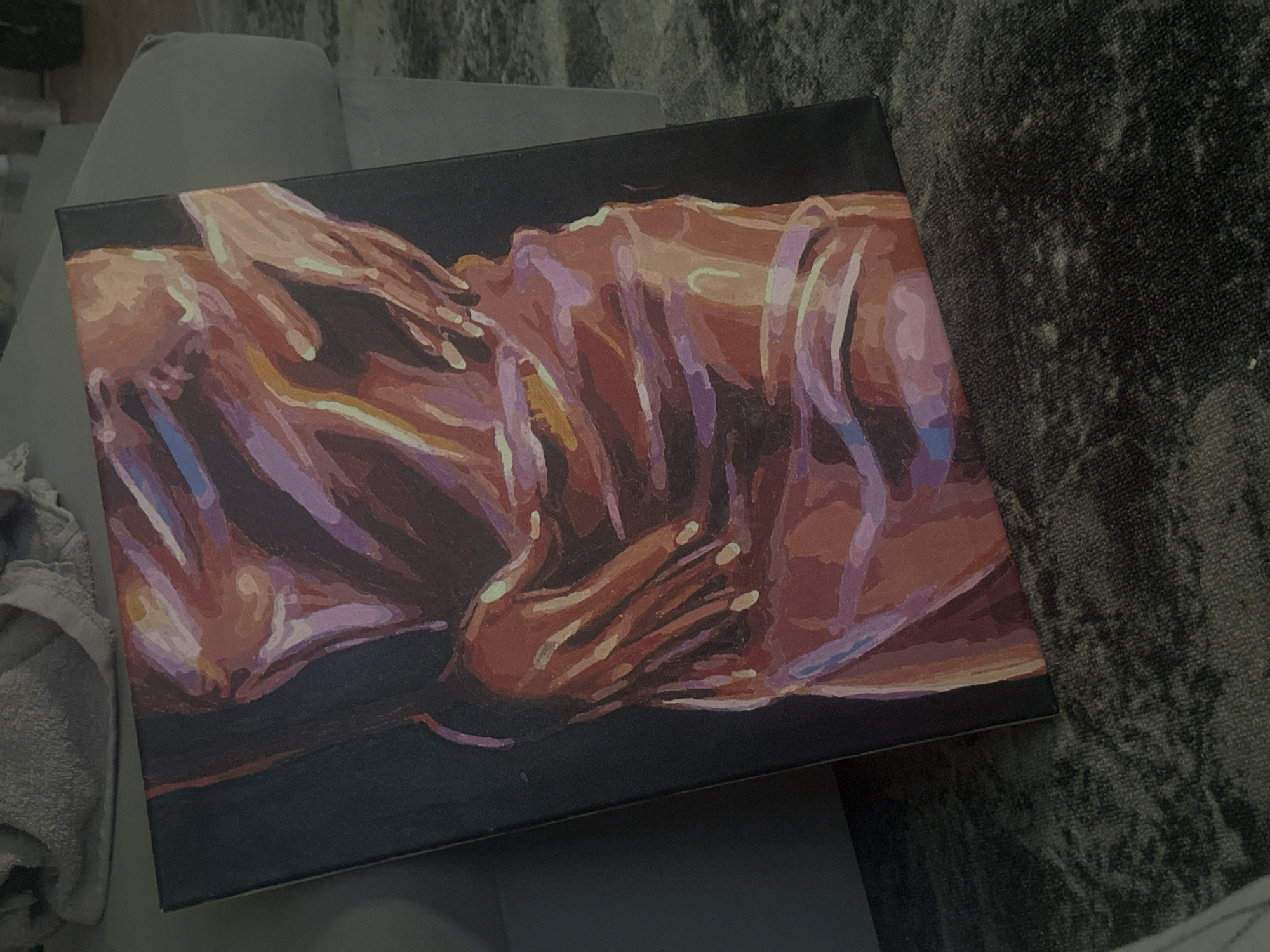 The image shows a painting on a canvas, likely depicting a human form or figures. The canvas is rectangular with a dark background. The figures are rendered in shades of orange, red, and purple. The artwork seems to be an abstract or stylized representation of a human subject. The painting rests on what appears to be a soft, possibly upholstered surface with a gray color. There are also visible textures of a carpet or rug in the background. There is no clear indication of the location in the city of Высокий, Russia.