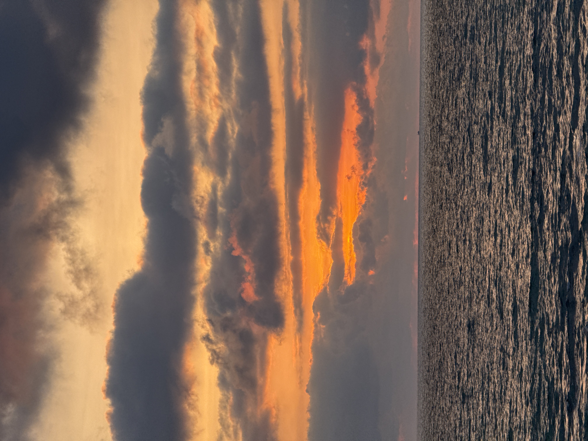 The image depicts a view of the sea at sunset from Tel Aviv, Israel. The sky is filled with dark clouds, some illuminated by streaks of orange and yellow light. The sea stretches to the horizon, with the surface rippled by waves. The horizon is distinct, separating the sea from the sky.