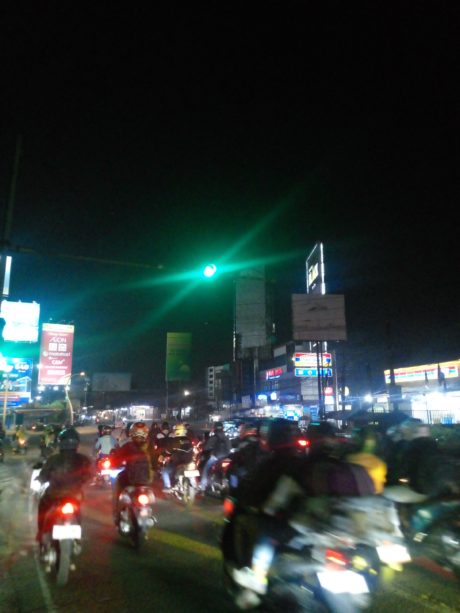 The image captures a nighttime street scene in Bitung Jaya, Indonesia, likely during a traffic jam. A prominent green traffic light is illuminated in the upper center of the frame, casting a glow over the surrounding area. A dense crowd of motorcyclists occupies the road, with many vehicles in motion, indicated by motion blur. The motorcyclists are varied in their attire, with some wearing helmets and others not. Several businesses and billboards are visible along the street, including what appear to be advertisements for "AEON" and "Matahari," suggesting a commercial area. The overall ambiance is one of a busy urban night, with the lights of vehicles and businesses creating a dynamic and somewhat chaotic atmosphere.