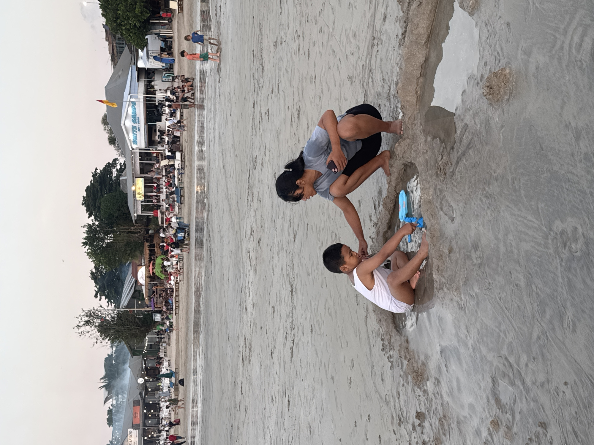 The image depicts a bustling beach scene at Kuta Selatan, Indonesia, likely during late afternoon or early evening given the soft lighting and overcast sky. In the foreground, a young boy wearing a white tank top is playing in the wet sand, holding a blue toy. Nearby, a young woman in a gray t-shirt and black leggings is crouched down, looking at her phone, with her feet in the water's edge.
The background reveals a lively beach with numerous people seated at tables and walking along the shore. Several structures, likely restaurants or shops, line the beach, adorned with colorful umbrellas and string lights. Smoke rises from some of the establishments, suggesting food preparation. Further down the beach, more people are visible, some standing and others seated. A red and yellow flag flies from a building. The sand is wet and reflects the dim light, indicating the tide is either going out or has recently come in.
Visible text includes "WHITE SANDS" and "EST - 1994" on one of the buildings. Another sign appears to read "SEAFOOD." The overall mood is relaxed and convivial, with people enjoying the beach and its amenities.