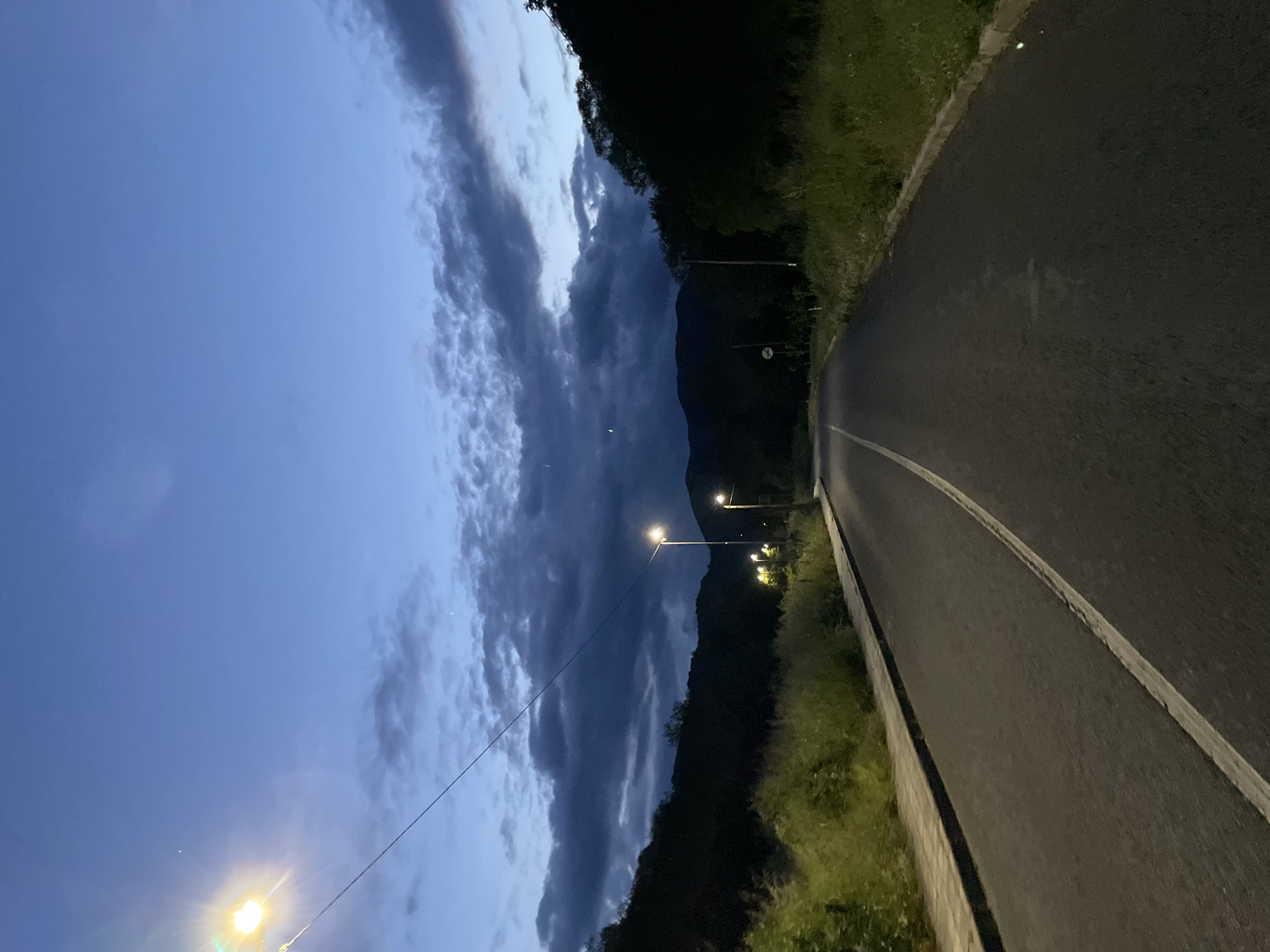 A low-angle, wide shot depicts a street, likely a road running through or near Zlatograd, Bulgaria, at dusk or early night. The road runs at a slight angle, top right to bottom left. The sky is visible on the left, transitioning from light blue at the horizon to darker blue and gray above. A long line of clouds stretches vertically along the sky. The road is dark asphalt with a painted white line along its left edge, and the land to the right is covered with grass and some foliage. Several street lamps illuminate the right side of the road, and a mountain is vaguely visible in the background. A power line is visible extending diagonally from the lower left to the center.