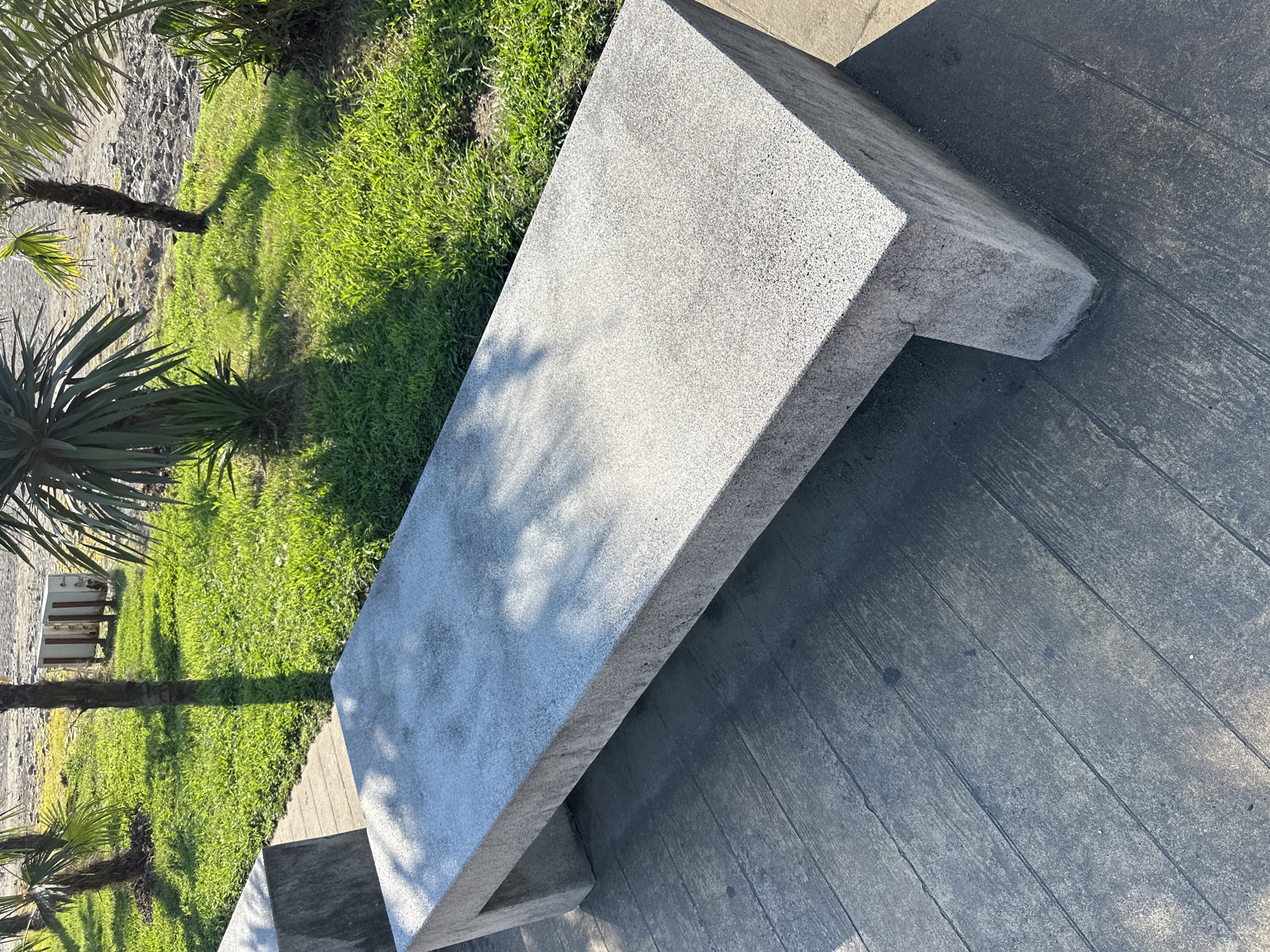 An eye-level, medium shot captures a long, modern, light gray speckled concrete bench situated on a dark gray, wood-plank-patterned concrete walkway. The bench is positioned at an angle, extending from the bottom left corner towards the center right of the frame. Sunlight casts dappled shadows from unseen trees onto the bench and the walkway. To the left of the bench, partially visible, is another similar concrete bench. Behind the walkway and benches, a vibrant green lawn is illuminated by bright sunlight. In the background, several palm trees and other greenery are visible, with a sandy or pebbly area and some distant structures suggesting a coastal or park environment. The scene appears to be in the city of Batumi, Georgia, given the presence of palm trees and the general ambiance of a seaside resort area. There are no people or animals present in the image.