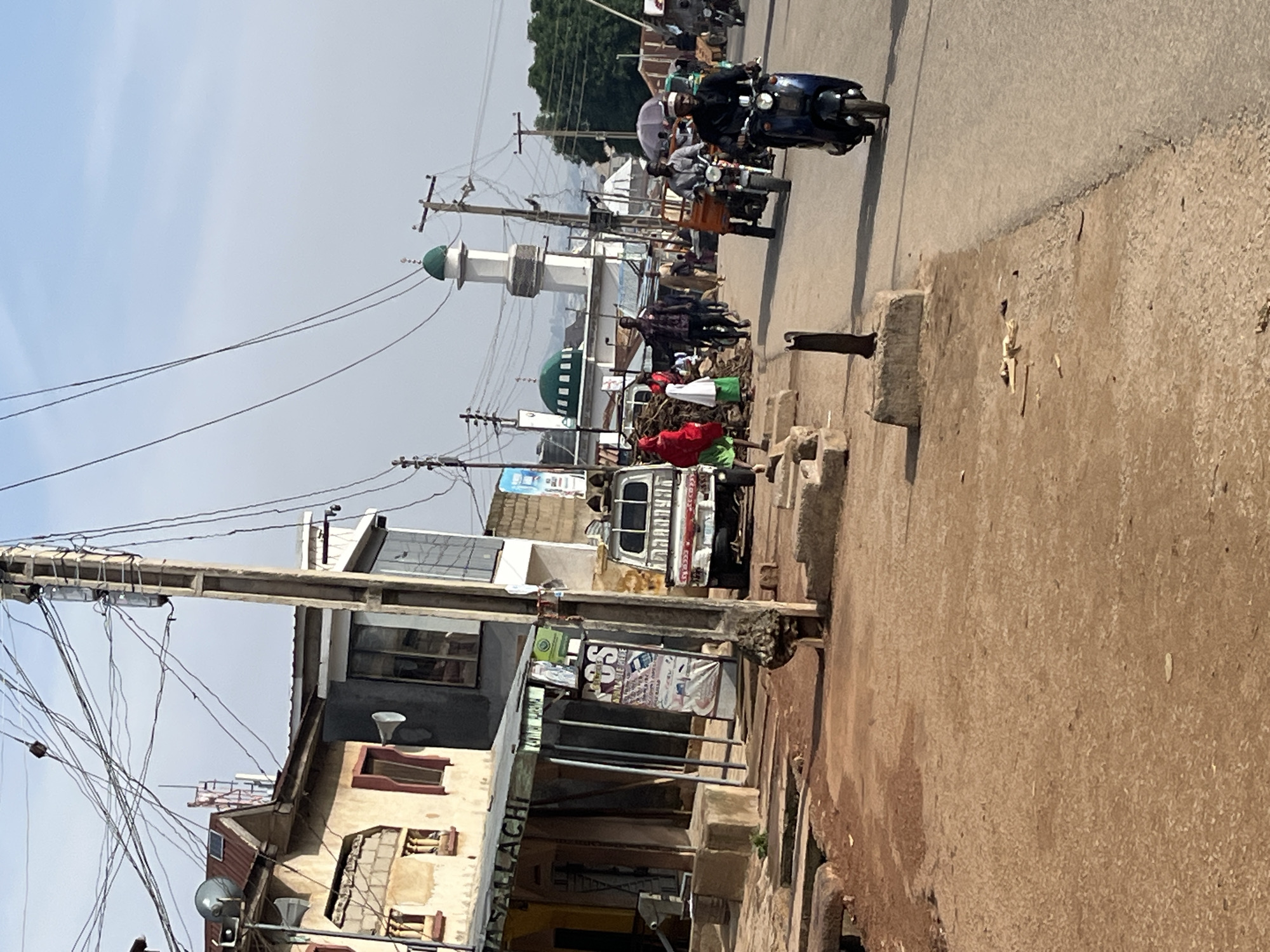 This is a street scene in Jos, Nigeria, featuring a bustling urban environment with a mosque in the background. The foreground shows a dirt road with some people walking and riding motorcycles. Several buildings line the street, some appearing to be shops with awnings. Electrical wires are prominently strung overhead, supported by utility poles. The weather appears to be sunny with a clear sky. In the distance, the distinctive dome and minaret of a mosque are visible, indicating a significant landmark within the city. The overall impression is one of everyday life and activity in a Nigerian city.