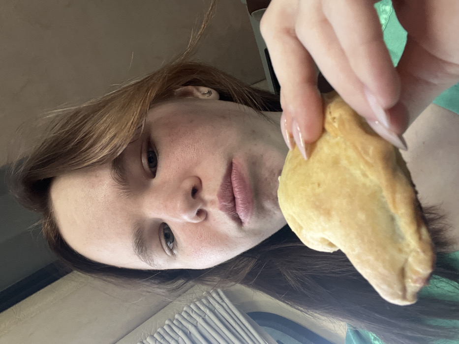 The image depicts a young female indoors in Yurkovo, Russia. Her face, viewed from a low upward angle, shows a light complexion with some facial redness and prominent brown hair with lighter highlights. A small earring is visible in her proper right ear. Her lips are slightly parted, suggesting an ongoing or imminent action of eating. She is holding a light golden-brown, irregularly shaped pastry in her proper right hand. Her fingernails are long, oval-shaped, and appear to have a natural or clear polish. She is wearing a green garment. The background consists of a blurry, light-colored ribbed panel or vent on the left and an otherwise indistinct, warm neutral-toned interior, consistent with an enclosed space.