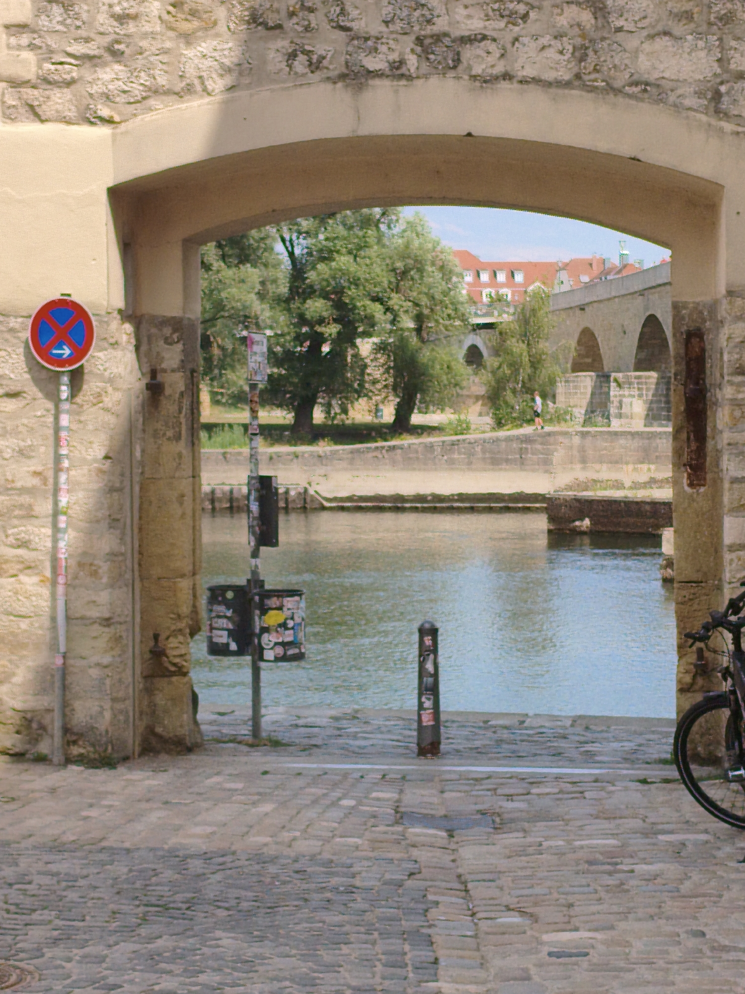 The image shows an archway in Regensburg, Germany, looking out onto a body of water and a bridge. On the left, a no parking sign with an arrow pointing to the right is mounted on a pole covered in stickers. A trash can, also covered in stickers, is located near the water's edge. The ground is paved with cobblestones. A bicycle is partially visible on the right side of the frame, parked near the archway. In the background, a man is walking along the water's edge with a bridge and buildings visible beyond him. The scene is illuminated by natural light, suggesting it is daytime.