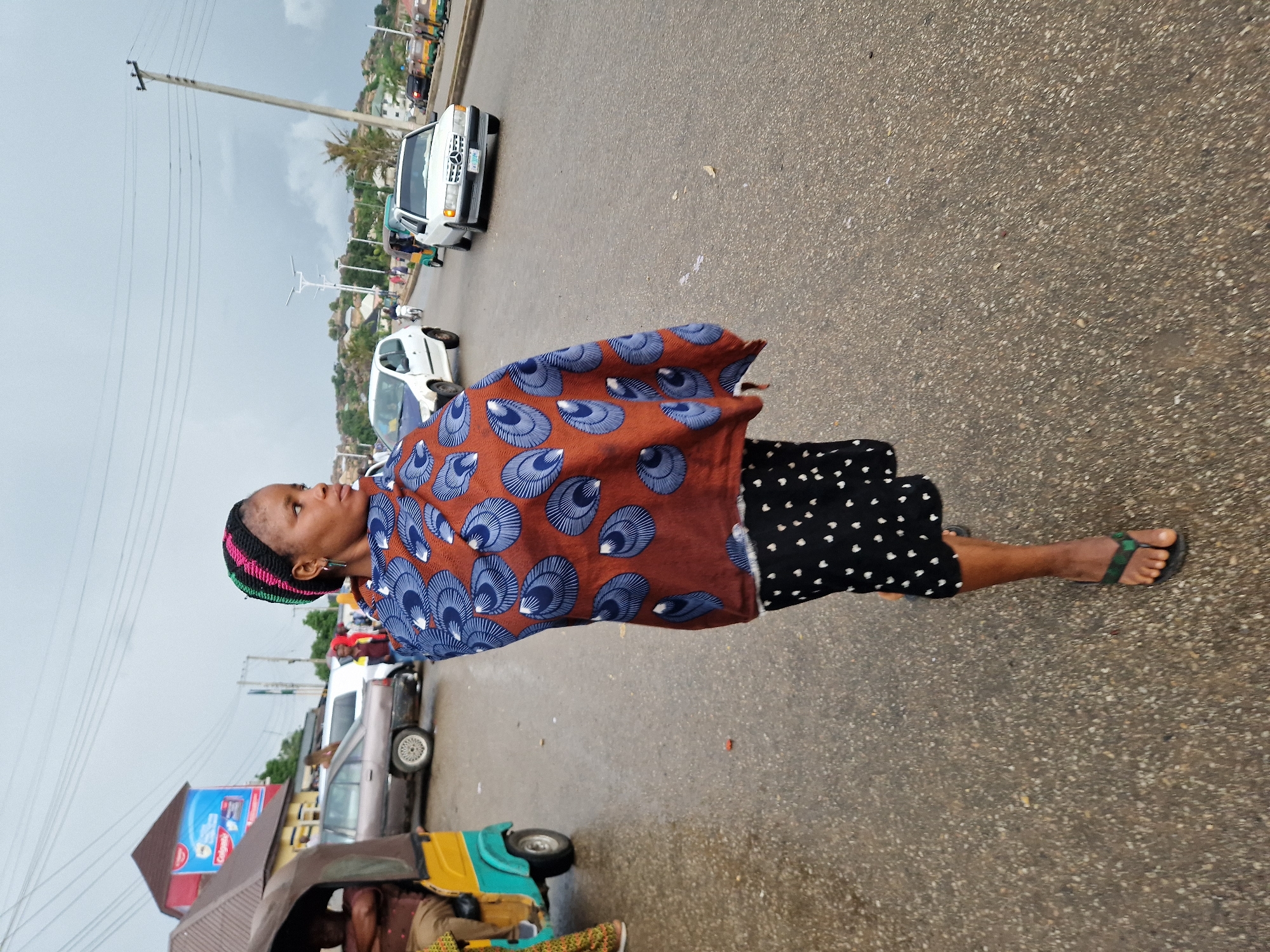 A woman stands on a wet asphalt road, looking to her right. She wears a colorful, braided headband, a brown and blue patterned wrap draped over her shoulders, and a black skirt with white polka dots. Her feet are clad in dark flip-flops. Behind her, traffic moves along the road, including cars, a white SUV, and a yellow and green three-wheeled auto-rickshaw. The scene appears to be in Kabong, Nigeria, suggested by the presence of a "Colgate" billboard and other visual cues characteristic of the region. The sky is overcast and the road surface is damp, indicating recent or ongoing precipitation.