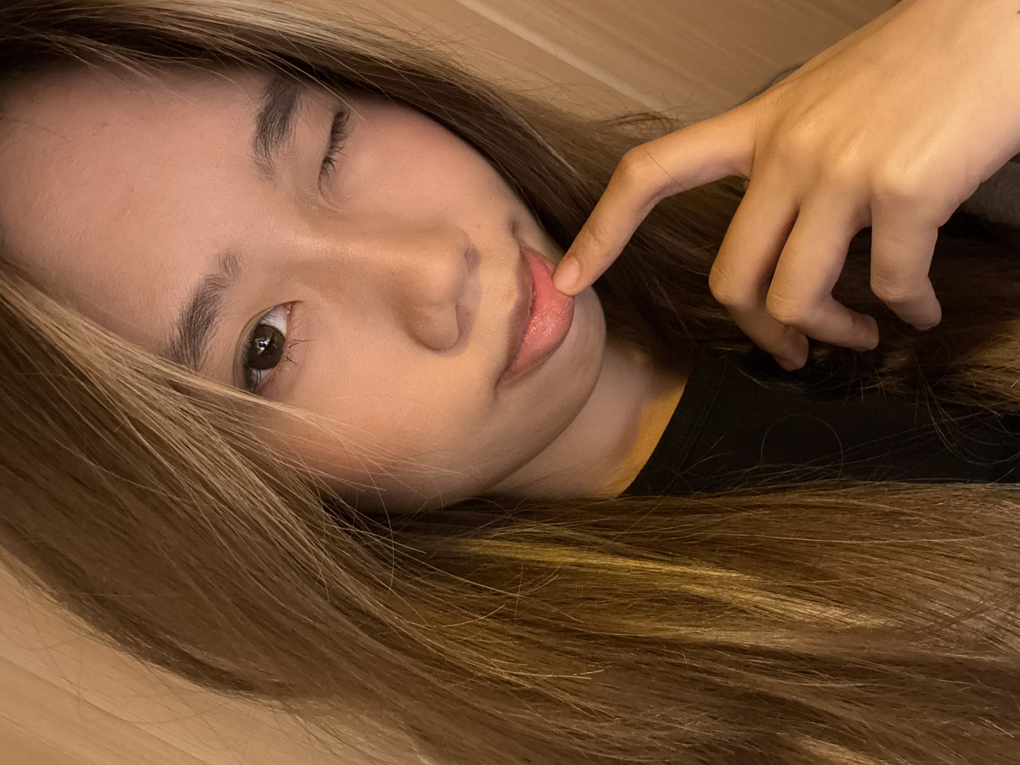 A woman lies down indoors. She has light brown hair with blonde highlights and is wearing a black shirt. Her index finger touches her lower lip. The location is impossible to determine definitively from the image alone.