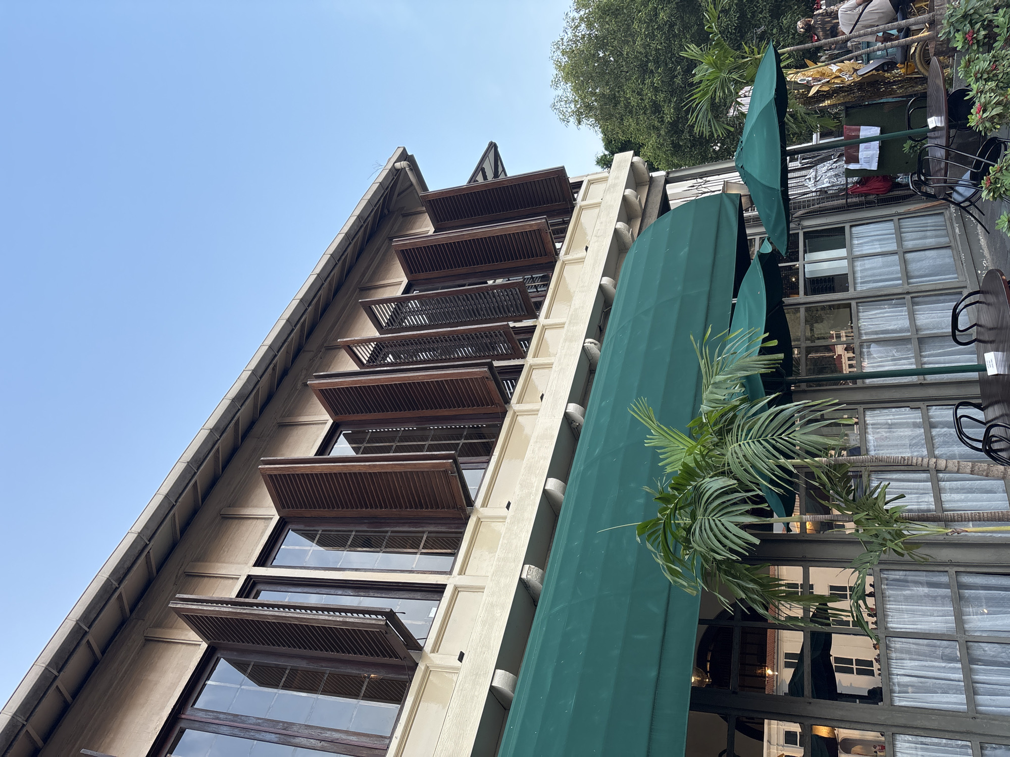 The image shows the exterior of a building, likely a cafe or restaurant, in West Jakarta, Indonesia. The building features a light-colored facade with multiple large windows on the upper floor, each fitted with dark wooden shutters that are partially open. A prominent green awning extends from the building's lower level, covering outdoor seating areas. Two green umbrellas are visible, providing shade for outdoor tables. Several potted palm trees are placed in front of the building, adding to the tropical ambiance. In the background, lush green trees and a clear blue sky are visible. A few people are present in the background, suggesting a public space. The overall scene depicts a serene and inviting outdoor setting.
