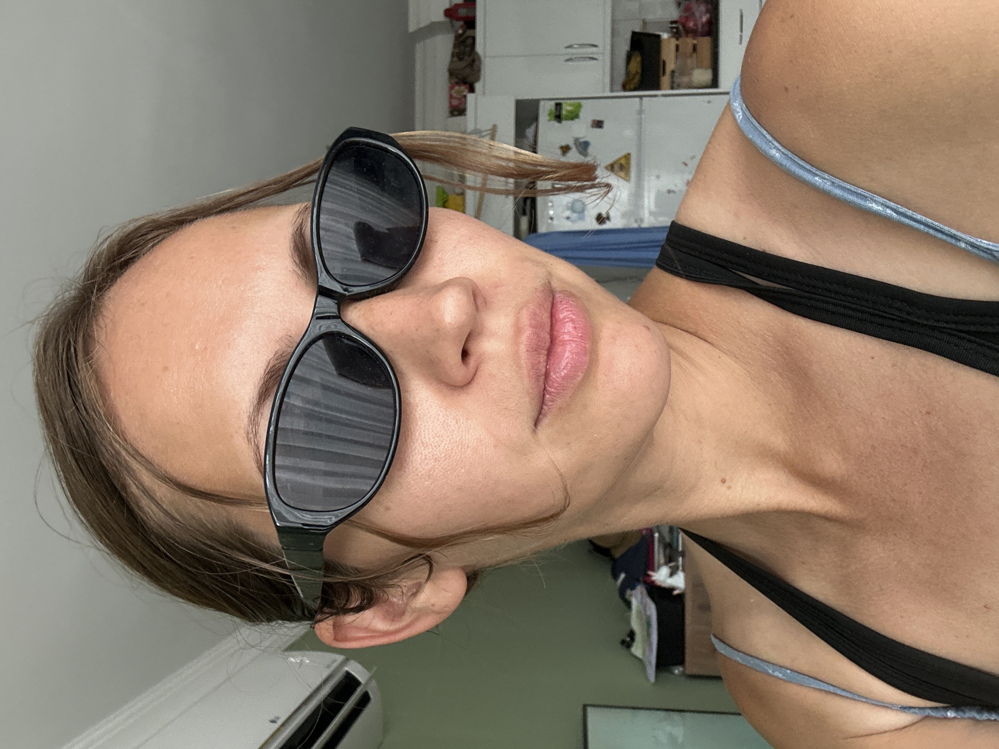 A woman is pictured from an unusual angle, wearing sunglasses. Her hair is light brown. She is wearing a black top with a blue strap. The location appears to be inside a building, potentially a residence, with a white wall and an air conditioning unit visible in the background. The scene is in Unknown, Türkiye.