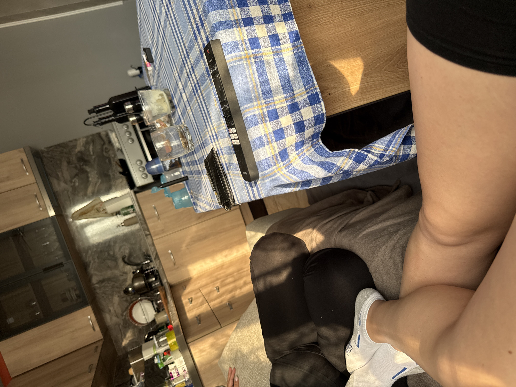 The image presents an indoor scene from a first-person perspective, likely a combined kitchen and dining area in a home in Rogosh, Bulgaria. In the immediate foreground, the bare legs and feet of a person are visible, suggesting they are seated on a grey blanket or cushion. The person wears black shorts or underwear, and white ankle socks with blue stripes.
A light wooden table is positioned to the left, draped with a blue and white checkered tablecloth. On the tablecloth, a black remote control is prominently placed, with the brand "PHILIPS" clearly visible on its surface. Other items on the table include a dark bottle, possibly wine, alongside another lighter bottle and two glasses, one of which appears to have some residue.
Beyond the table, a kitchen area features light-colored wooden cabinets and drawers. A reflective stainless steel countertop or splashback holds various kitchen items, including a coffee machine or similar appliance, other bottles, and utensils. Strong natural sunlight streams into the room, casting distinct shadows on the table and kitchen cabinets, indicating it is daytime, possibly morning or late afternoon. The overall atmosphere suggests a casual, lived-in domestic setting.