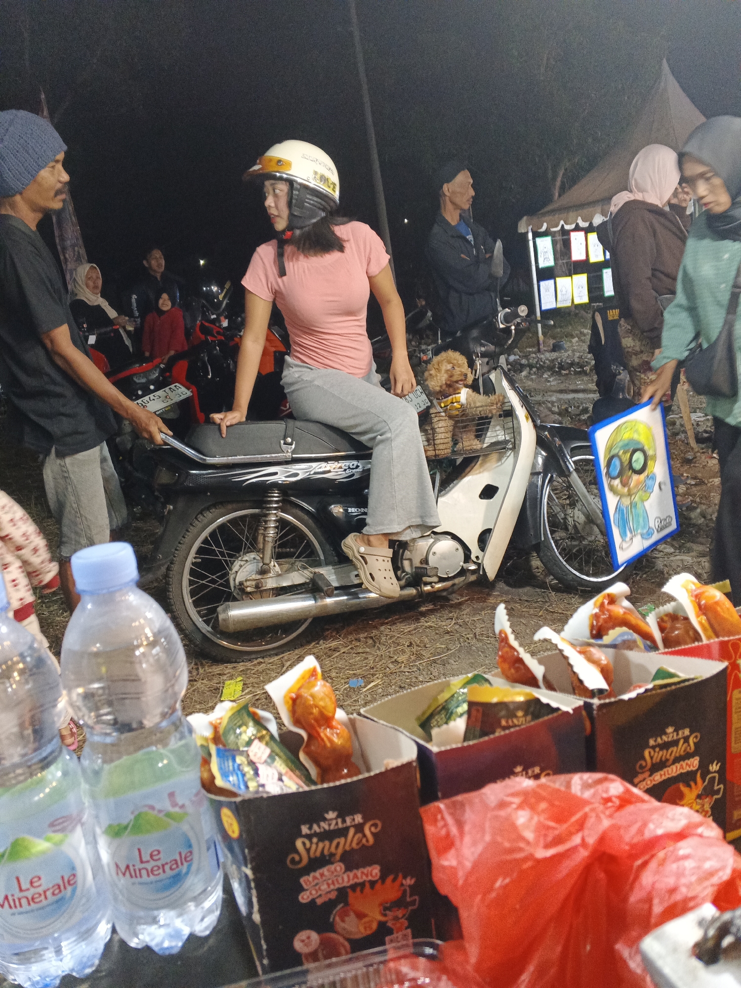 The image depicts a nighttime outdoor scene, likely a street market or event, in Sukabumi, Indonesia. In the foreground, two bottles of "Le Minerale" water are prominently displayed. Behind them, several open cardboard boxes contain what appear to be packaged food items, with some red packaging visible. A bright red plastic bag is also in the foreground, partially obscuring the food boxes.
The central focus of the image is a black motorcycle, on which a woman wearing a pink t-shirt and a white helmet is seated. She is looking to her right, and her left hand rests on the motorcycle's seat. To her left, a man wearing a dark t-shirt and a blue beanie stands beside the motorcycle, holding onto the rear passenger grip. A small, light brown poodle is visible in a basket attached to the front of the motorcycle.
In the background, several other people are present, some standing and some seated on motorcycles. There is a tent or stall structure with framed pictures or posters displayed on it. The overall environment appears to be somewhat cluttered, with loose debris on the ground. The lighting suggests artificial illumination, common for nighttime events.